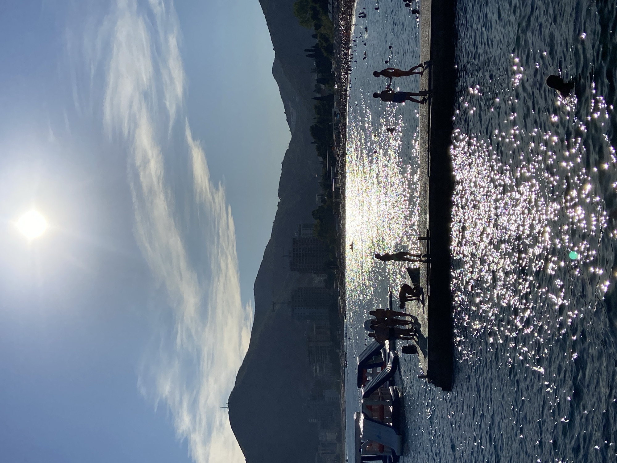A bright, sunny day at a beach in Budva, Montenegro is depicted. The sun is high in the sky, casting a strong glare on the water, creating a shimmering effect. In the foreground, a pier extends into the sea, with several people silhouetted against the bright water. Some individuals are standing on the pier, while others appear to be preparing to enter the water or are already in it. To the left of the pier, a section of an inflatable water park is visible.
In the background, a wide expanse of the Adriatic Sea stretches out, dotted with swimmers. The coastline is lined with buildings, some of which are under construction, and lush green hills rise behind the city. The overall scene conveys a lively atmosphere of a popular tourist destination on a clear, warm day.
