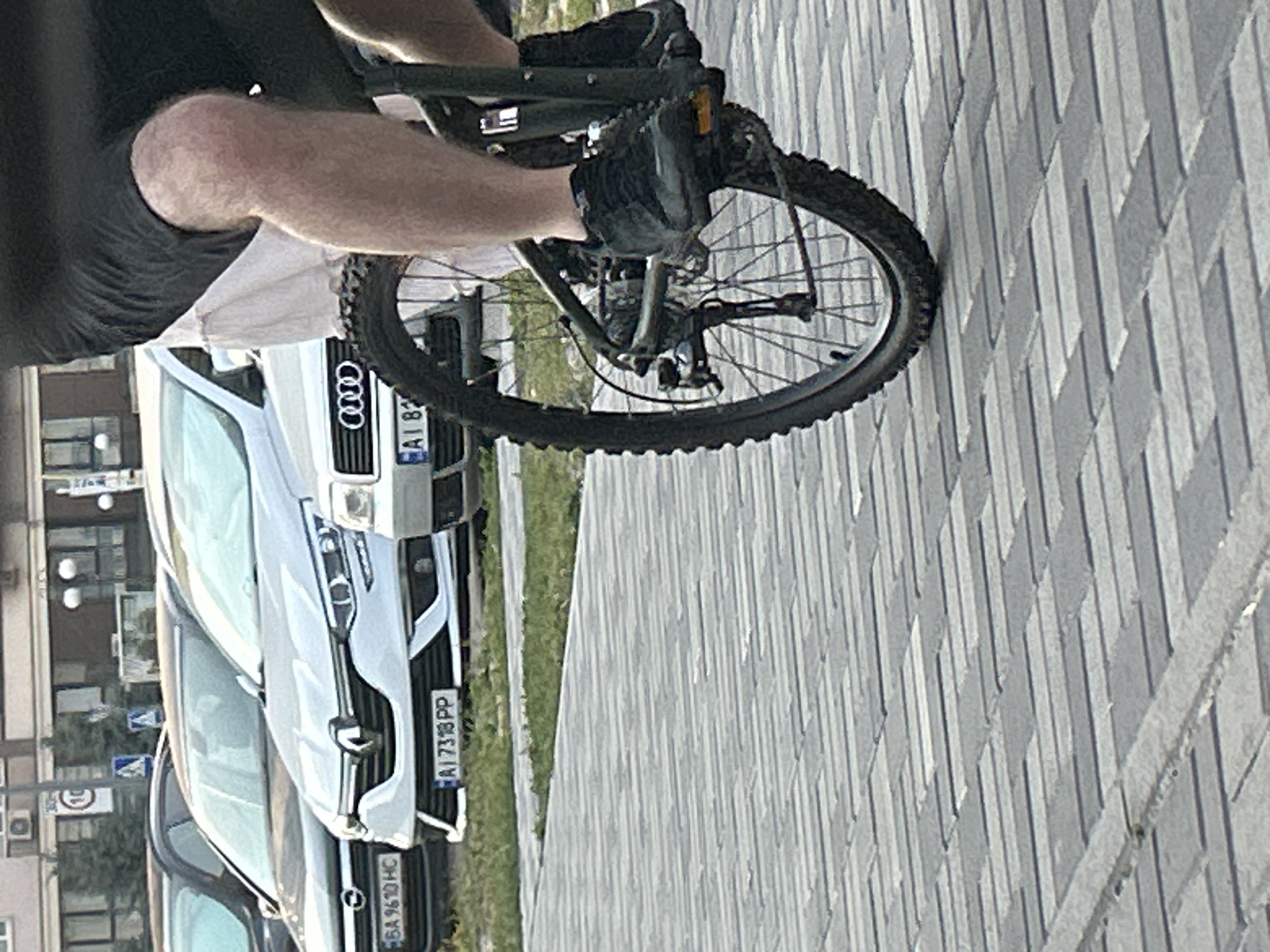 An individual, seen from the knees down, rides a bicycle on a paved walkway. They are wearing black shorts and black sneakers. The bicycle has a dark green frame and a knobby tire. In the background, several cars are parked. One car, a silver Renault, has a license plate reading "AI 7318 PP." Another car to its left, possibly an Opel, has a license plate reading "BA 9610 HC." An Audi is also visible, with its distinctive four-ring logo. Buildings and trees are visible in the far background. The scene appears to be a street or public area in Brovary, Ukraine.