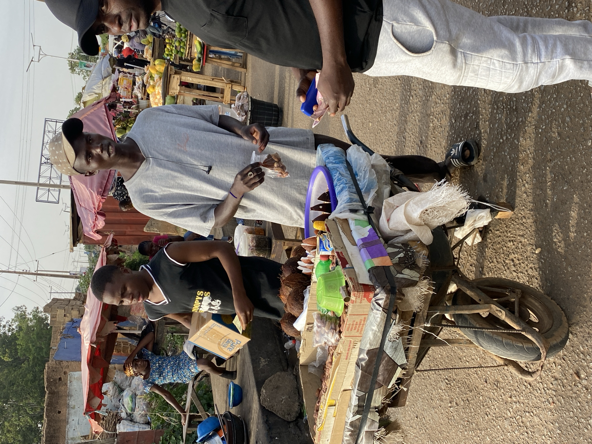 In Bukuru, Nigeria, a street scene captures a makeshift vendor's bicycle cart laden with assorted items, including coconuts, packaged goods, and plastic containers. The cart is situated on a paved street, flanked by what appears to be a bustling marketplace. Two men stand beside the cart; one is handling a small plastic bag containing food, while the other examines a blue object. A third man is positioned at the back of the cart, handling a box. Behind the cart, there are structures, one partially covered with a pink and white canopy, suggestive of market stalls. A woman is seen attending to something near the rear stalls. The environment seems typical of a busy urban market setting.