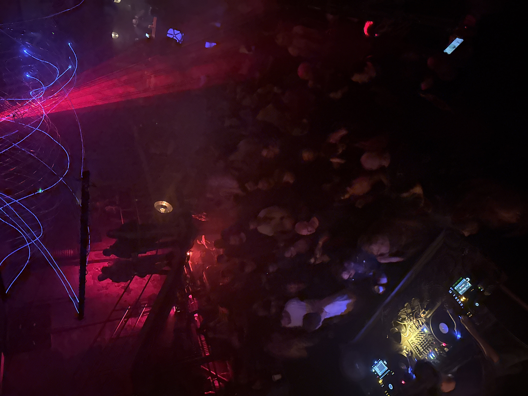 The image depicts a dimly lit nightclub, likely in Moscow, Russia, given the lighting and architecture style. A crowd of people is present, their faces indistinct due to the low light. Lighting includes laser beams in blue and red, casting an eerie glow on the crowd. A DJ booth with turntables and other equipment is visible on the right. People are likely dancing and socializing.