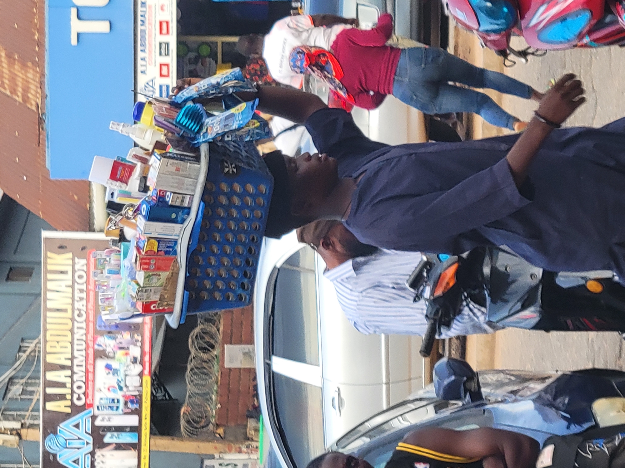 A man carries a blue plastic basket filled with various items on his head, seemingly a street vendor, in what appears to be a bustling urban area. The background includes buildings with signage, possibly indicating businesses. Other people are visible, including one in jeans and a red top and another in a striped shirt. Vehicles, including a car and a motorbike, are also present. The sign A.I.A ABDULMALIK COMMUNICATION is visible in the background. The location is the city of Jos, Nigeria.