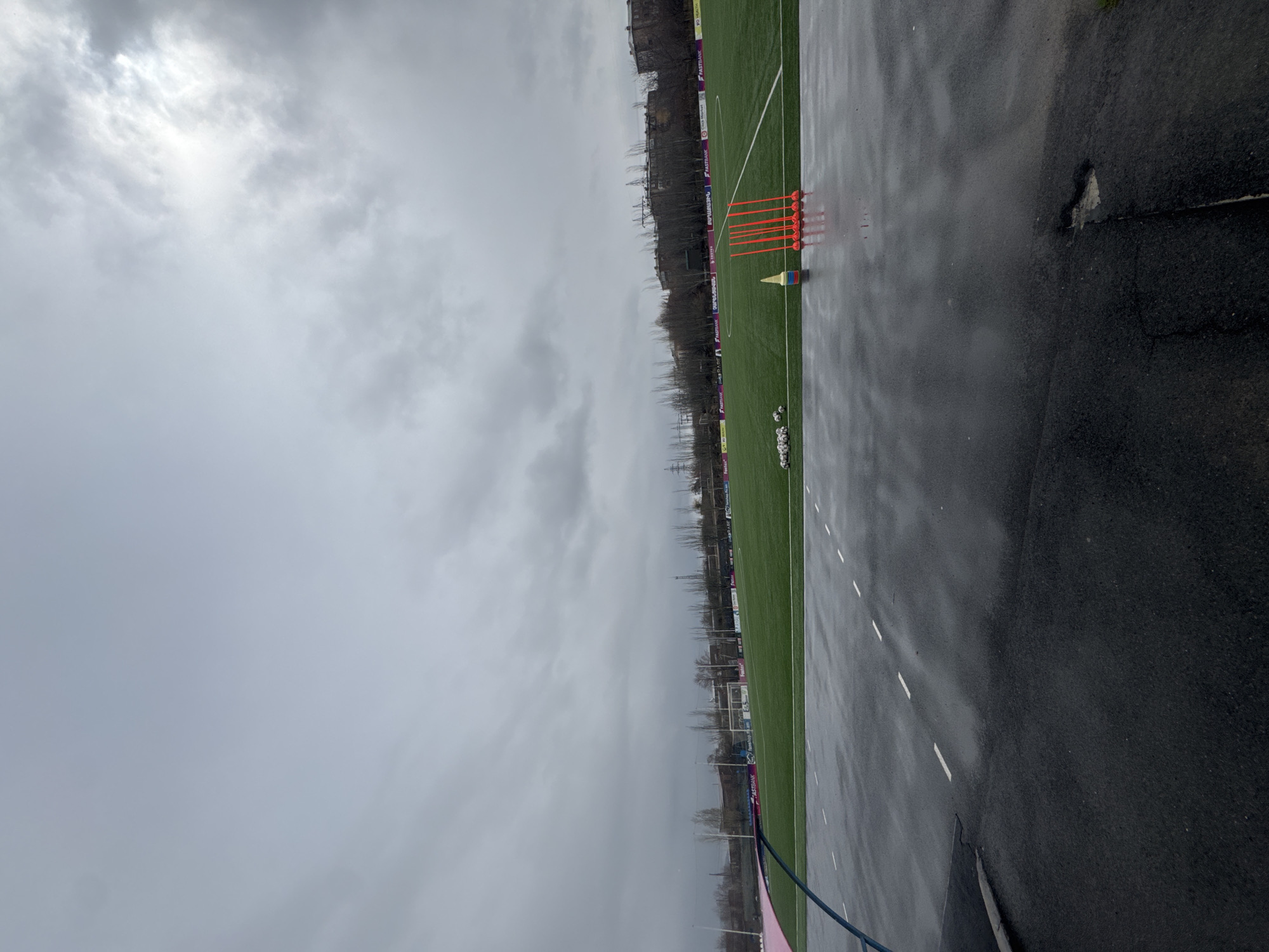 The image shows a view of a soccer field and surrounding area in Charentsavan, Armenia on a cloudy, overcast day. The field is green artificial turf, with white lines marking the boundaries. Orange cones and other training equipment are placed on the field. A stack of soccer balls is visible. Advertising boards line the side of the field. The foreground is a wet, paved surface, possibly a running track or walkway. Beyond the field, there are trees and buildings, and utility poles are visible in the distance. The sky is a uniform grey, indicating overcast weather.