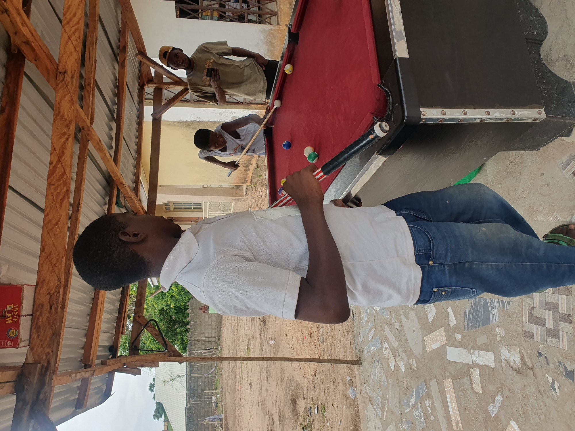 In Lokoja, Nigeria, three young men are playing pool in an outdoor, open-air setting. The scene appears to be under a makeshift shelter with a corrugated metal roof supported by wooden beams. The ground is a mix of dirt and decorative tiles. One person, wearing a white polo shirt and blue jeans, is in the foreground, leaning over the pool table and holding a cue stick. He is focused on the game, with a determined expression.
In the background, two other young men are visible. One, wearing a beige t-shirt and a yellow baseball cap, is looking at his phone. The other, wearing a grey t-shirt, is also near the pool table, watching the game. The pool table itself has a red felt surface and is scattered with several billiard balls. The lighting suggests it is daytime, likely late morning or afternoon, with natural light illuminating the scene. The environment seems to be a casual recreational area, possibly a backyard or a community space. There is a red box with visible text on the left side of the image; "TASTY TOM" is legible on it, possibly indicating a food product. The overall atmosphere is relaxed and social, with the men engaged in a friendly game of pool.