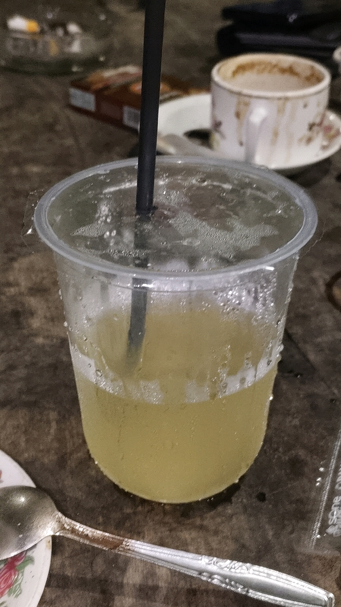 A close-up, vertical, eye-level shot shows a cold, light-yellow beverage in a clear plastic cup on a dark, textured surface, possibly a wooden table. The cup is filled to about three-quarters capacity, and condensation is visible on the exterior. A black straw protrudes from the center of the cup's lid, submerged in the liquid. In the background, a white teacup with a floral pattern sits on a saucer, containing a frothy, light-brown beverage, likely coffee. A pack of cigarettes is also partially visible. A silver spoon with an ornate handle lies in the foreground, partially obscuring a small white plate with a floral design. The overall scene suggests a casual setting, possibly a cafe or a home in Karangtalun, Indonesia.