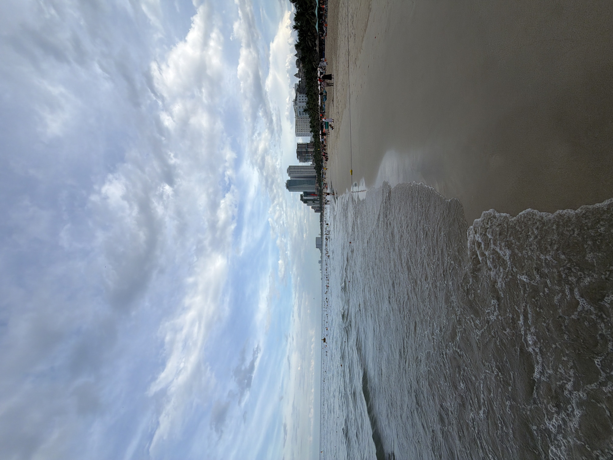 A panoramic view of a crowded beach in Da Nang, Vietnam, under a cloudy sky. The foreground features the gentle waves of the ocean lapping onto the sandy shore. In the midground, numerous people are spread across the beach, some swimming in the water and others relaxing on the sand. The background reveals a line of modern high-rise buildings and palm trees, indicating an urban coastal setting. The overall atmosphere is that of a popular tourist destination on a moderately overcast day.