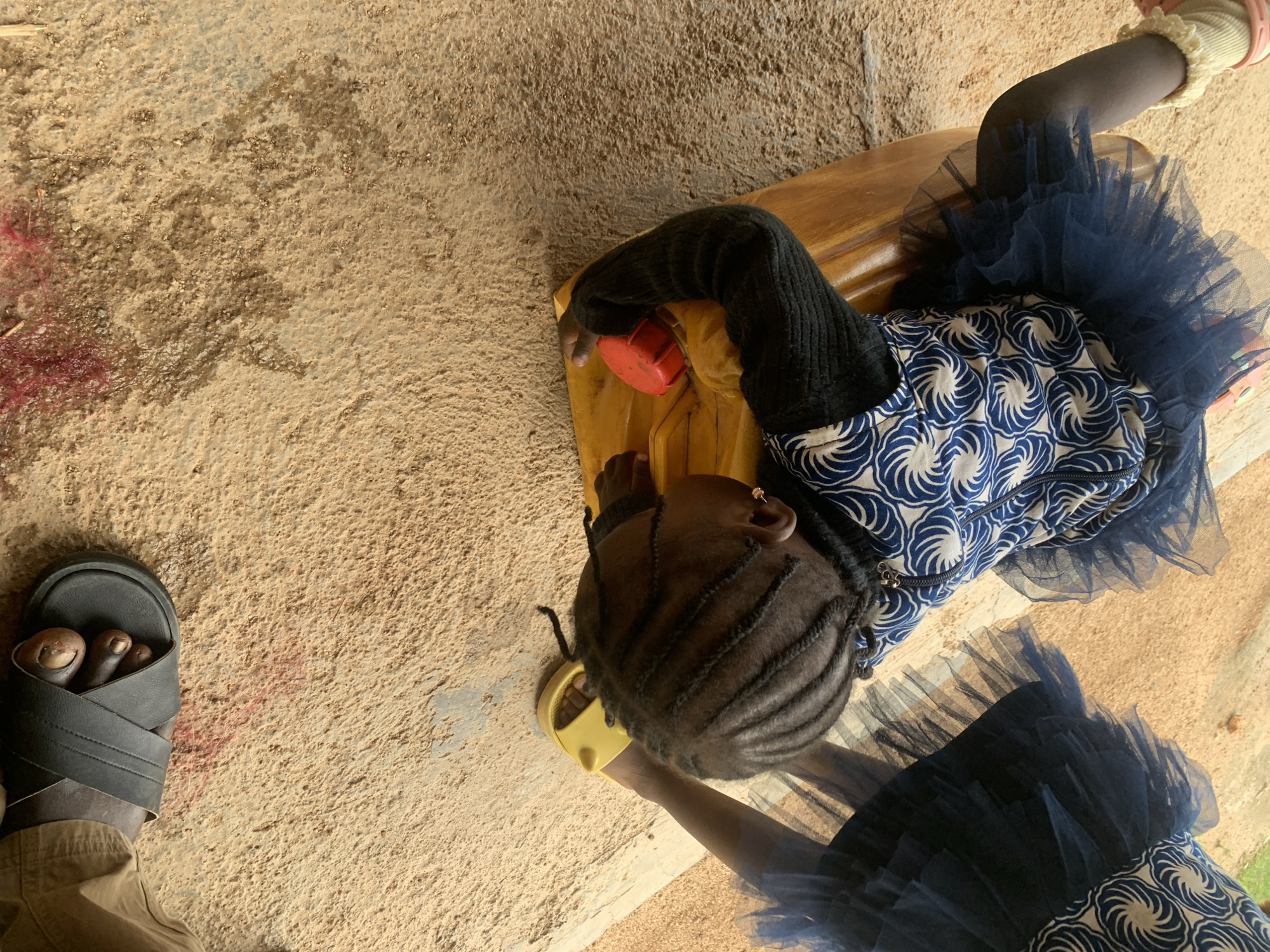 This image captures an overhead, slightly angled view of two young children on a sandy surface, likely outdoors in Jos, Nigeria. One child is wearing a black long-sleeved top and a blue and white patterned dress with a dark blue tutu skirt. This child is holding a bright red-capped yellow container with their right hand. Their hair is styled in braids, and they are wearing a gold hoop earring. The child is seated or leaning on a light brown wooden surface.
To the left of the first child, another child's foot is visible, wearing a black slide sandal. This child is also wearing tan-colored trousers. The sandy ground appears to have some reddish-pink stains on it, possibly from paint or dye, and some scattered debris. The overall lighting suggests it is daytime. No specific weather conditions are apparent. The scene appears casual, possibly a moment captured during play or an activity. There is no visible text in the image.