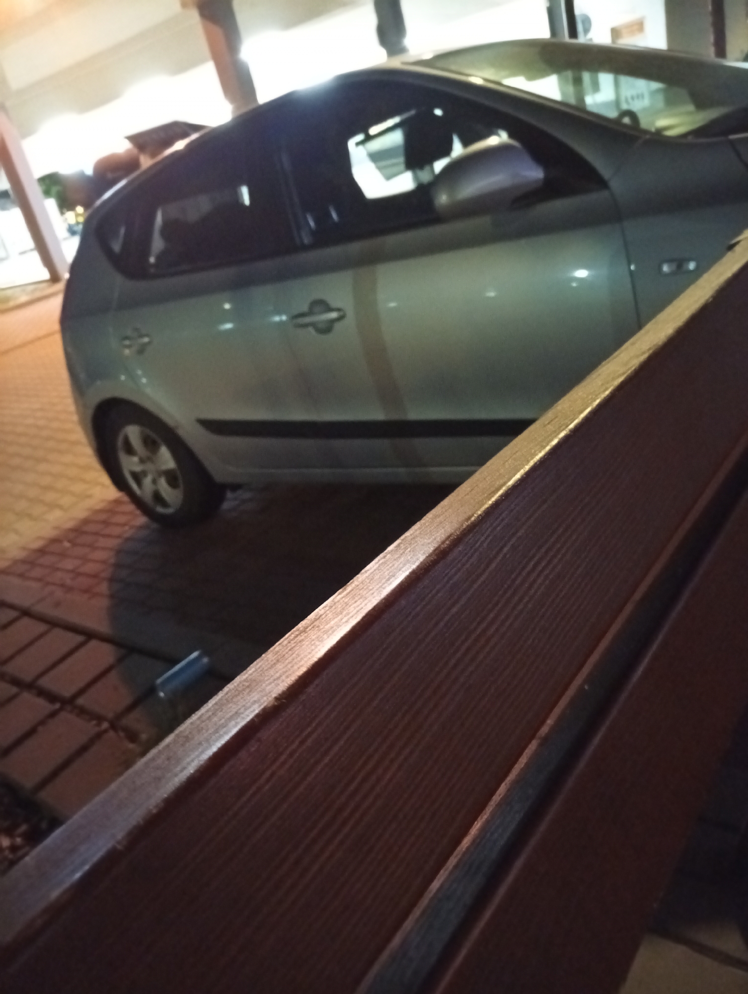 The image captures a side view of a light-colored hatchback car parked outdoors at night. The car's doors, windows, and wheel are visible. In the foreground, a wooden bench with a visible grain pattern is positioned at an angle, partially obscuring the car. The ground beneath the car appears to be paved with bricks. The lighting suggests an urban environment, possibly a street or parking area, with artificial lights casting reflections on the car's surface. There are no people visible in the image. The location is identified as Tišnov, Czechia.