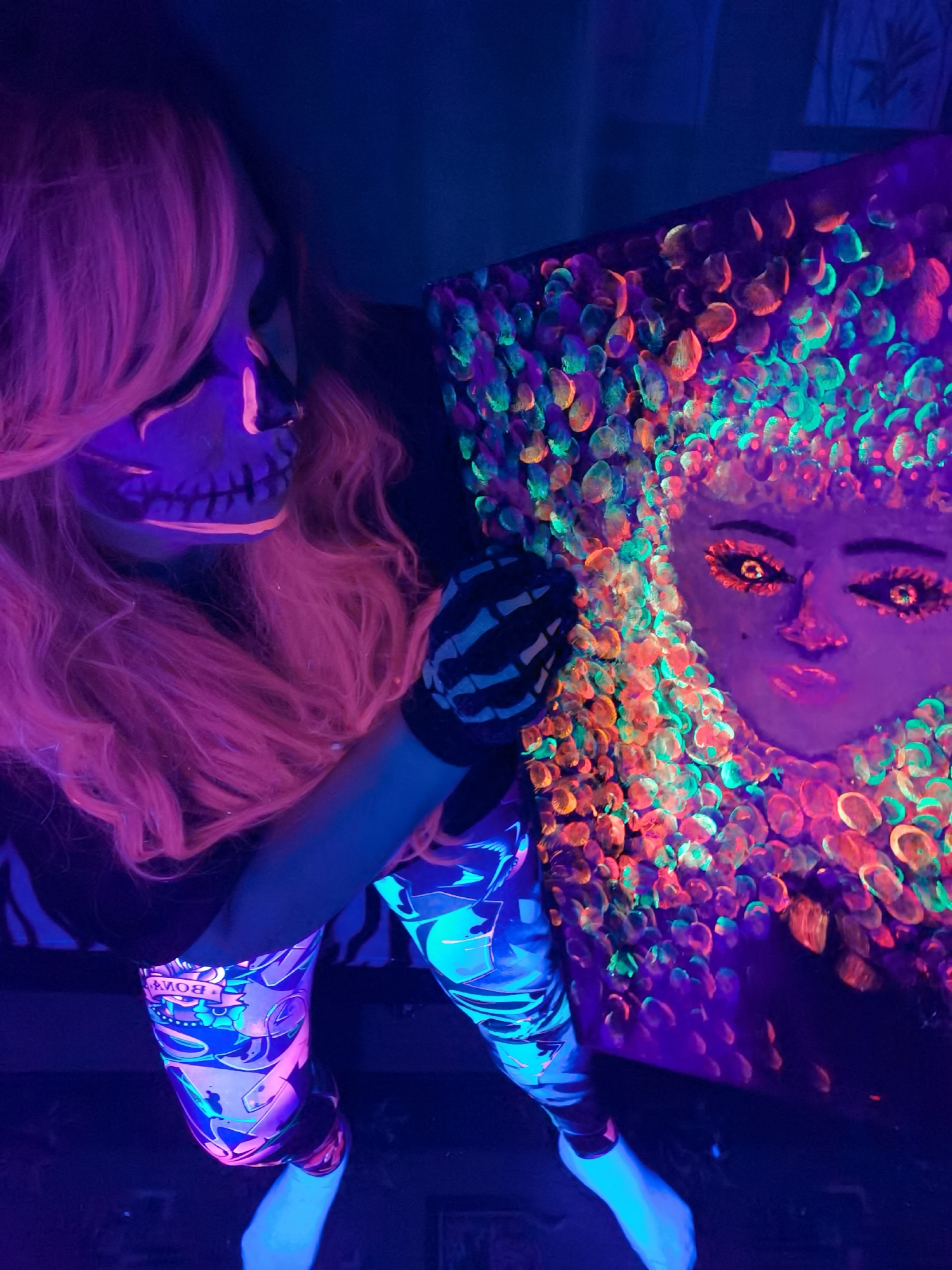 This media file captures a person displaying a piece of artwork in a blacklight setting, likely indoors in Chaikovsky, Russia. The individual, possibly female, has long, flowing pink hair and an elaborate skull-like face paint design that glows vibrantly in fluorescent orange and pink under the UV light. They wear a black shirt and distinctive leggings covered in a glowing, multi-colored pattern that includes shapes and text; the word "BOMB" is visible in reverse on the left leg. A glove with a skeletal design is worn on their right hand.
The person is holding a square canvas painting that is the central focus. The painting features a stylized face, depicted in a subdued purple under the blacklight, with highly expressive, intensely glowing eyes that display intricate detail. Surrounding the face is a dense, textured aura or "hair" composed of numerous small, circular or oval marks, possibly fingerprints, that fluoresce brightly in shades of green, orange, and pink. The overall scene is dark, allowing the fluorescent elements of the person's attire, face paint, and the artwork to stand out dramatically, creating an artistic and somewhat mysterious atmosphere characteristic of a blacklight art show, performance, or photo shoot.