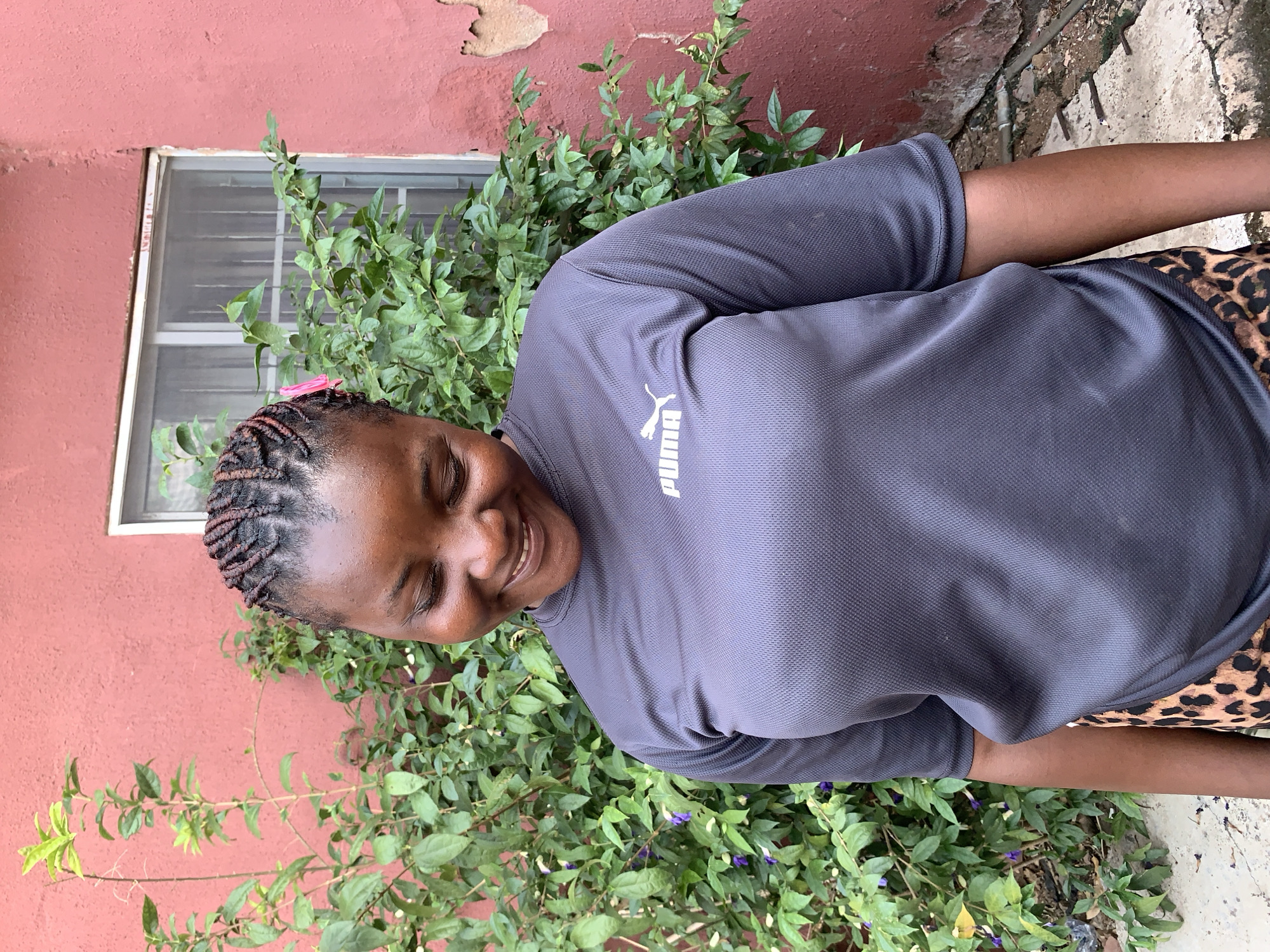 A portrait photograph, taken outdoors in Jos, Nigeria, features a young adult Black woman smiling broadly with her eyes gently closed. She is positioned sideways, against a backdrop of a reddish-orange textured wall and verdant green plants. The woman wears a dark grey short-sleeved t-shirt, clearly displaying a white "PUMA" logo on the chest, and leopard print bottoms. Her hair is neatly styled in small braids, and a small pink hair clip adorns her hair near her forehead.
The setting suggests a bright, clear daytime, possibly in a garden or yard. Behind the woman, a white-framed window with horizontal bars is partially visible on the worn reddish-orange wall. Lush green shrubbery with small purple flowers grows abundantly along the base of the wall. The ground beneath her is a cracked concrete surface. The woman's joyful expression, characterized by her wide smile and relaxed eyes, conveys happiness and contentment.