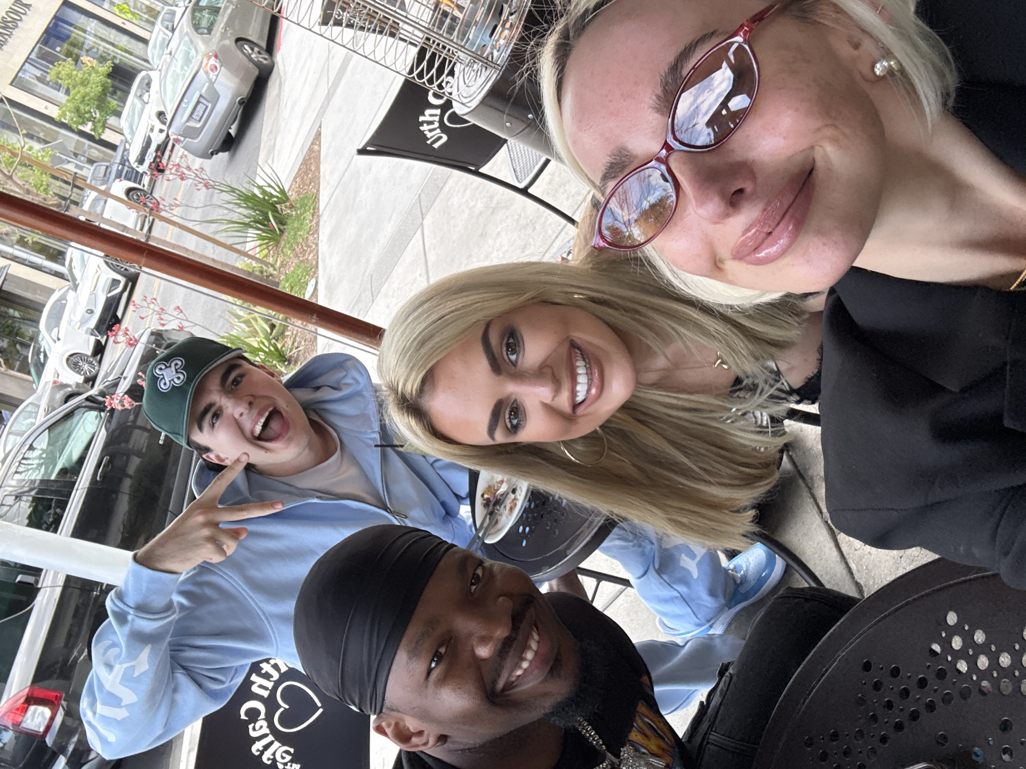 Four individuals are captured in an outdoor selfie setting in West Hollywood, USA. A woman wearing pink-rimmed sunglasses and a black jacket is positioned at the top right corner of the frame. Next to her, a blonde woman with gold hoop earrings smiles towards the camera. A young man in a light blue hoodie and green baseball cap flashes a peace sign. Completing the group, a man wearing a black head covering smiles from the lower center. They appear to be sitting at a table, with a partial view of a black umbrella and a table with a plate. The backdrop reveals parked cars and the cityscape of West Hollywood.
