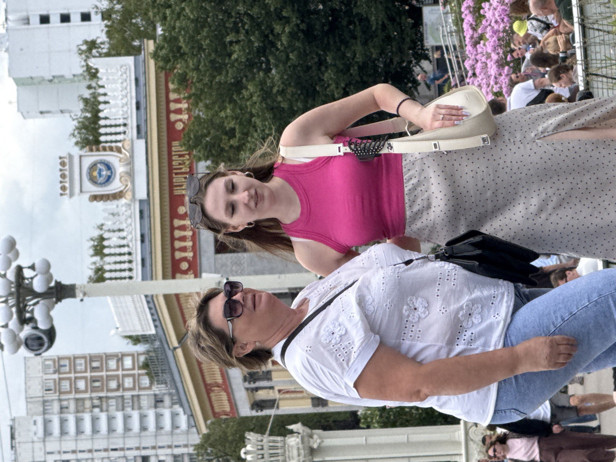 The image captures two women standing outdoors in front of a decorative building with a prominent clock tower. The architecture suggests a location within Moscow, Russia, possibly an exhibition center or park from the Soviet era. The woman on the left is older, wearing a white embroidered blouse and blue jeans, and carrying a black handbag. The woman on the right is younger, dressed in a bright pink tank top and a polka-dot skirt, with a light-colored shoulder bag. She has a ring on her left hand. In the background, a crowd of people can be seen, with lush green trees and a large expanse of pink flowers. The overall atmosphere is casual and summery.
