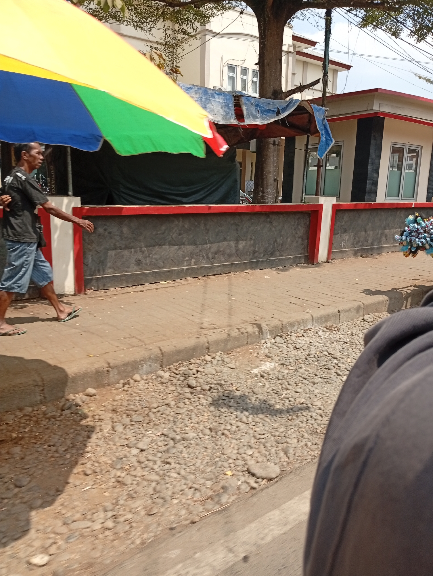 A man in a black t-shirt and blue shorts walks on a sidewalk. He is under a large, multicolored umbrella that is yellow, blue, and green. The sidewalk is made of interlocking bricks. A concrete curb separates the sidewalk from a gravelly area. In the background, there is a building with white walls and red trim. A large tree is also visible. To the right, a vendor is selling items, possibly balloons, in a bunch of blue and white. The overall scene suggests an outdoor market or street setting.