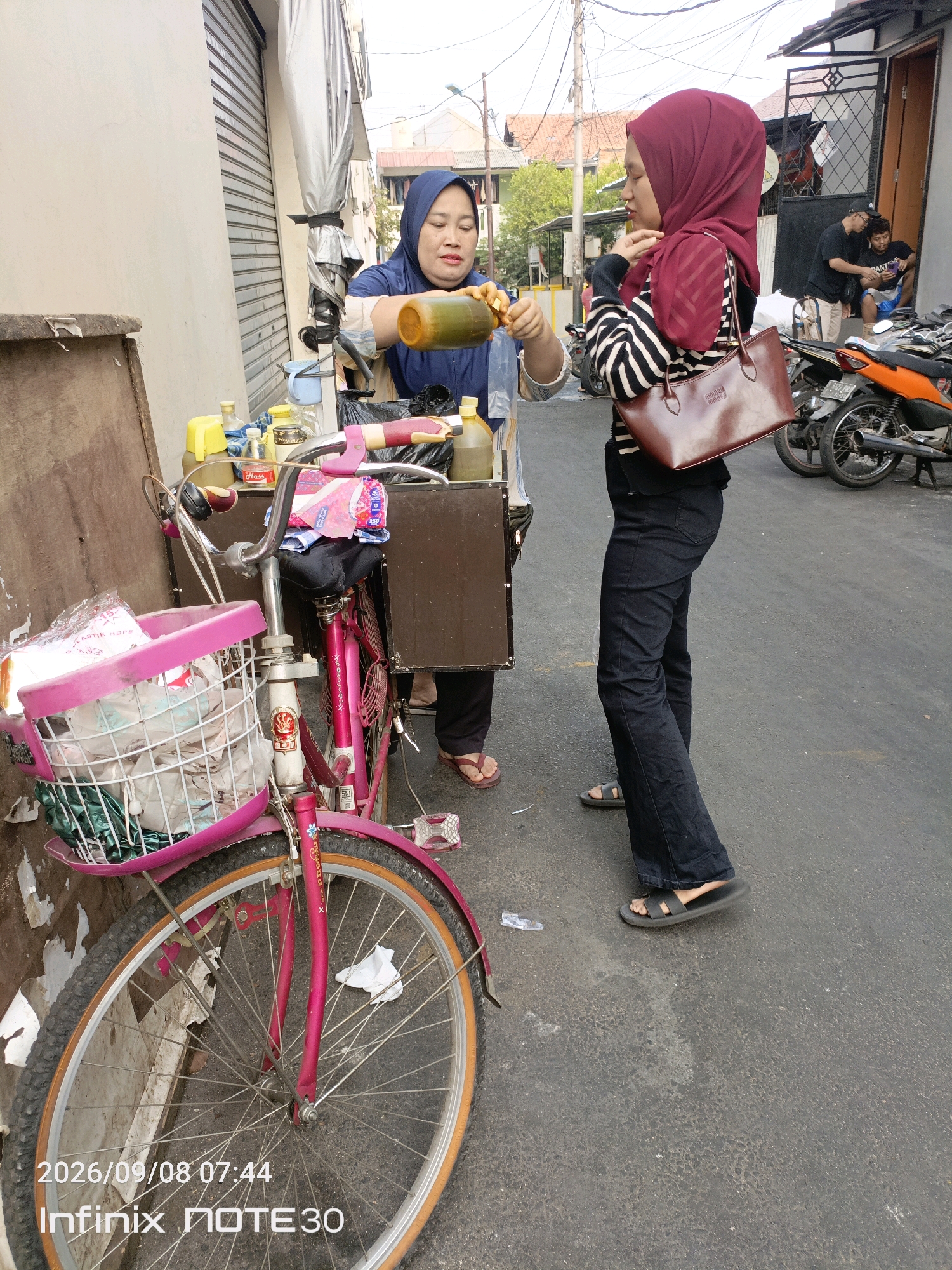 A street vendor in Central Jakarta, Indonesia, serves a customer from a cart attached to a pink bicycle. The vendor, wearing a blue headscarf and apron, is offering a food item from a large bottle of green liquid. The customer, in a maroon headscarf and striped shirt, holds a brown handbag and appears to be receiving the food. The background shows a street with buildings, parked motorcycles, and other people. The scene is captured with a date and time stamp of "2026/09/08 07:44" and "Infinix NOTE30".