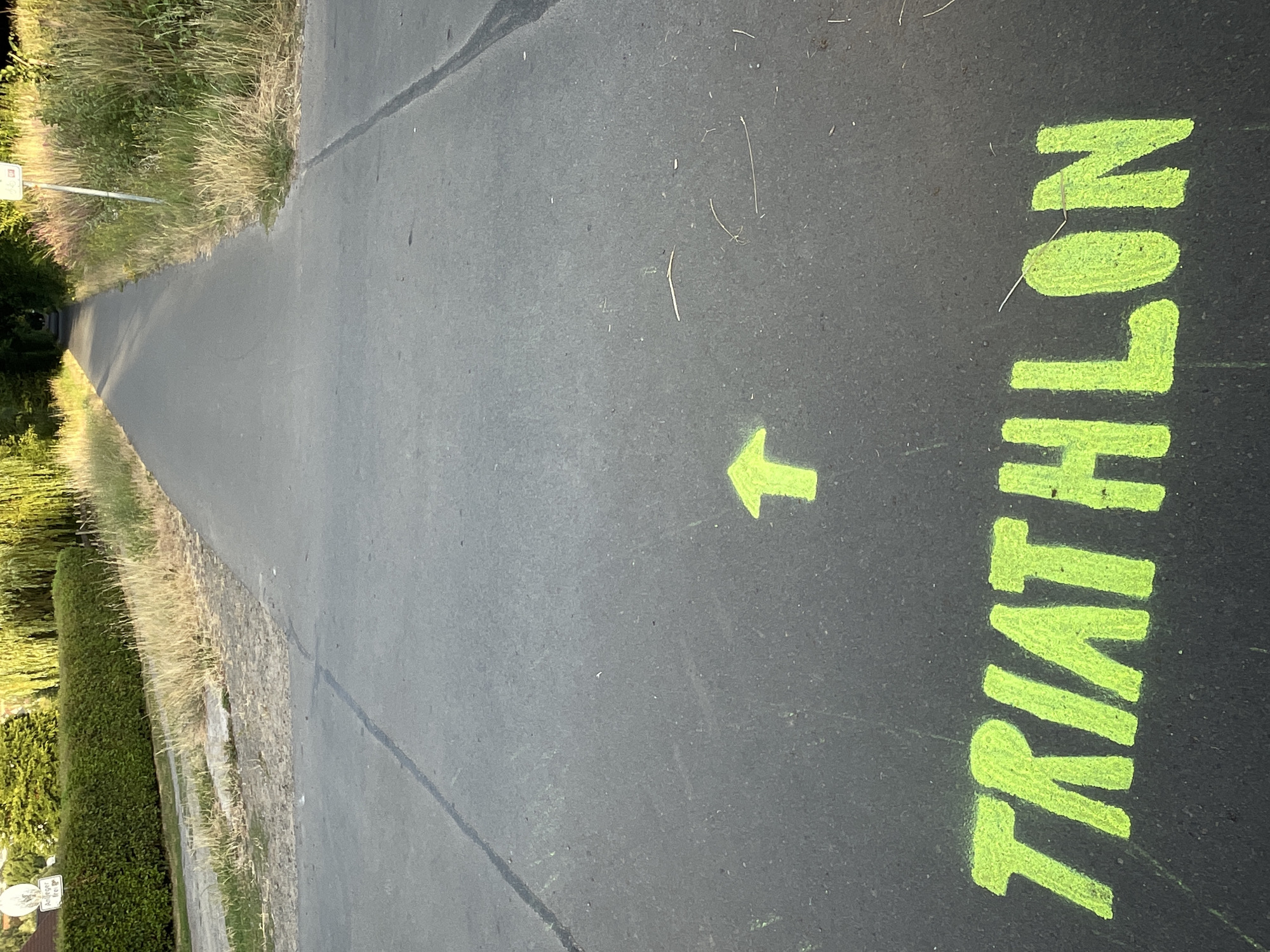 The image depicts a paved pathway with bright green chalk markings. The word "TRIATHLON" is spray-painted in capital letters on the asphalt in the foreground, accompanied by an upward-pointing arrow. The pathway stretches into the distance, lined with grass and trees on either side. A sign is visible on the right side of the path. The scene appears to be an outdoor course or event. It is located in Neukirchen, Germany.