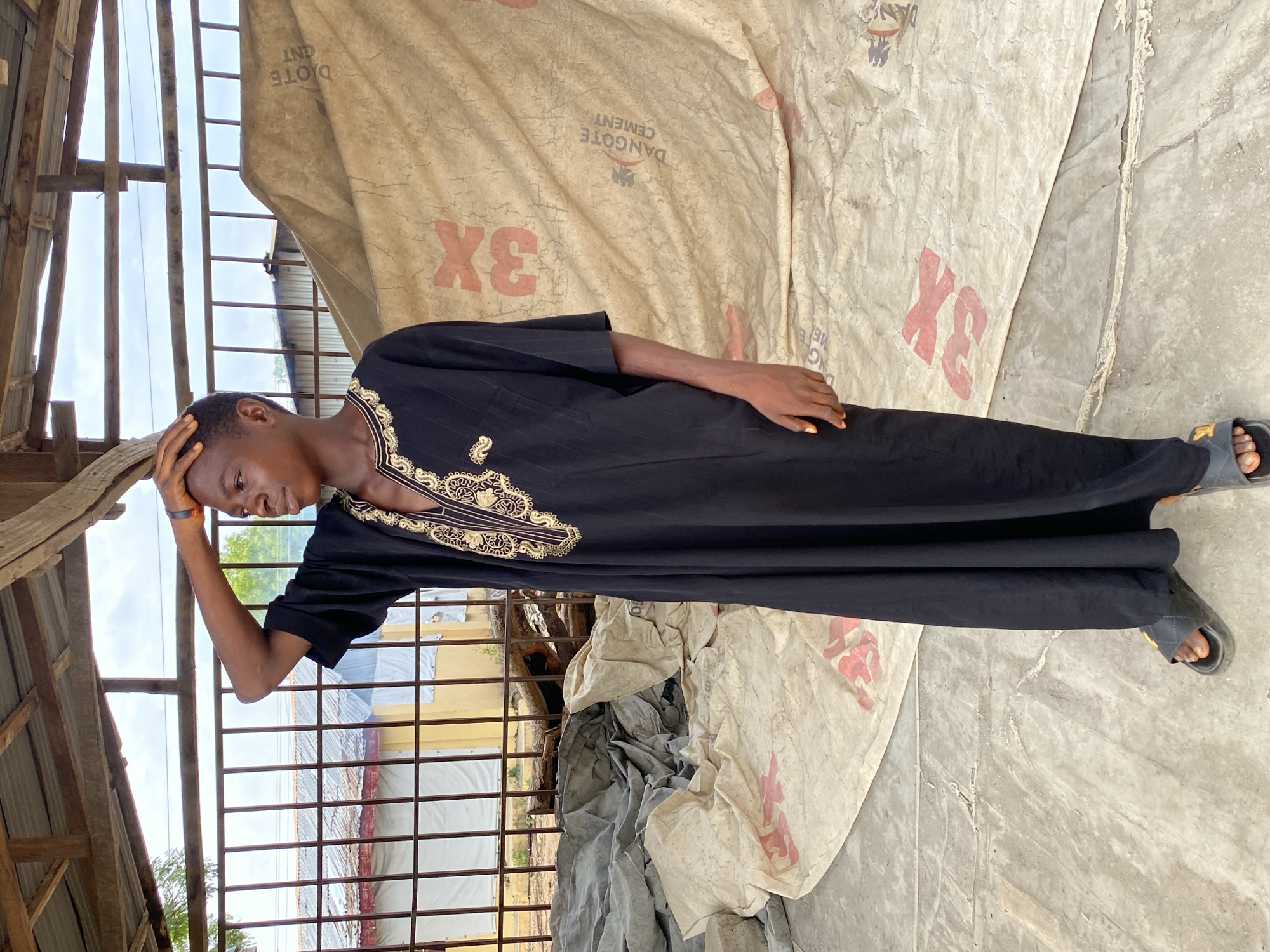 A young male, possibly in his late teens or early twenties, stands outdoors in Adankolo, Nigeria. He is dressed in a long, flowing black garment with intricate gold embroidery around the collar and chest, paired with black open-toed sandals. His left hand rests on his head, and he is looking down and slightly to his left, possibly in thought or weariness.
The setting appears to be a semi-enclosed or sheltered area, possibly under a makeshift structure. Above him, wooden beams and a metal grid-like structure are visible, with an overcast sky showing through. The background features a large, light-colored, crinkled material, which appears to be repurposed tarpaulin or heavy-duty canvas. This material has red text printed on it, including "DANGOTE CEMENT" and "3X," suggesting it might be from cement bags or similar industrial packaging. To the left, more of the grid structure and distant foliage can be seen. The ground beneath him is a light, uneven concrete or dirt surface. The overall impression is one of a utilitarian or construction-adjacent environment, likely during daytime under a cloudy sky.