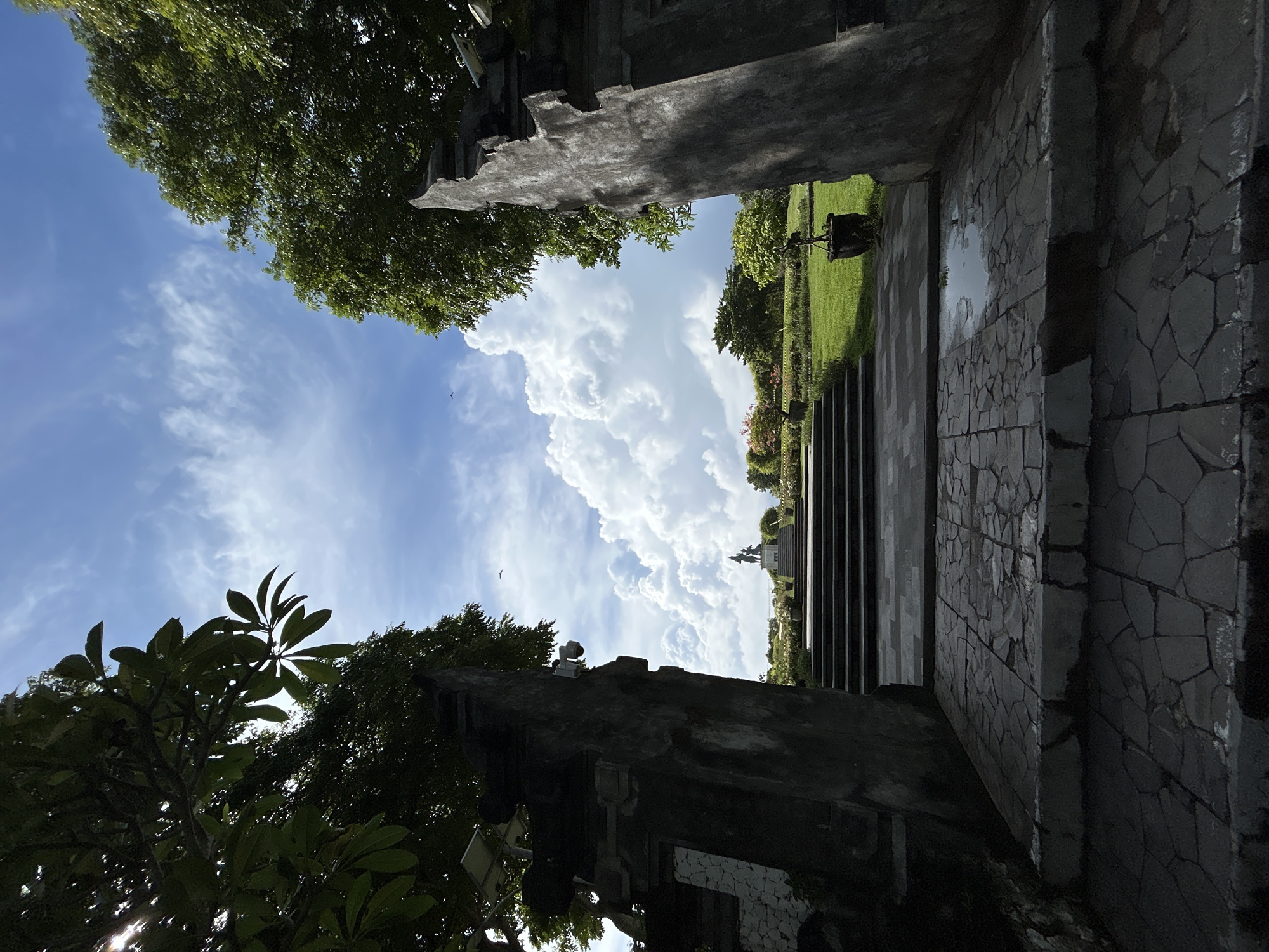 The image, oriented correctly, depicts a bright outdoor scene in Kuta Selatan, Indonesia, viewed from a shaded, dark stone structure looking out into an open area. In the immediate foreground, dark, rough-hewn stone walls and structures flank both sides of the frame, suggesting a gateway or an architectural opening. Dense green foliage from nearby trees is visible on the left, with some leaves silhouetted against the sky, and also in the lower right corner. A security camera is mounted on the upper part of the right-hand stone structure.
The central part of the image is dominated by a clear blue sky filled with large, fluffy white cumulus clouds, indicating bright daylight, likely mid-morning or early afternoon. A small, dark bird is visible flying in the sky. Below the sky, a lush, manicured green lawn extends into the midground. A wide set of dark stone steps leads downwards from the viewer's position into this grassy area. In the distance, at the far end of the garden, a light-colored statue or monument, possibly of a figure or deity, is visible. More trees and greenery frame the distant landscape.
No people are present, and no specific activity is taking place. The scene conveys a sense of tranquility and spaciousness, highlighting the natural beauty and traditional architectural elements under a bright, sunny sky with scattered clouds. No visible text is present.