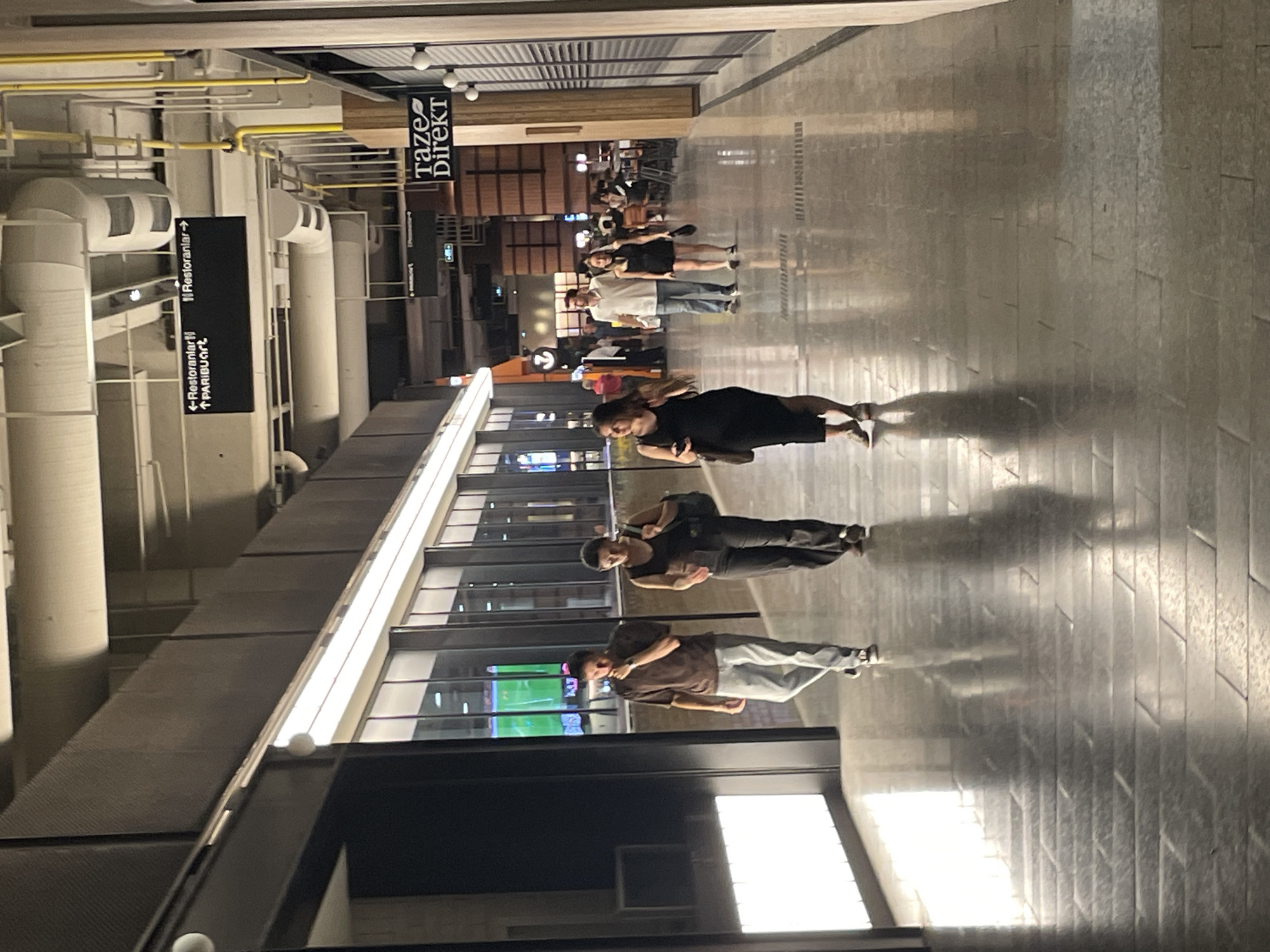 The image depicts a modern indoor walkway, likely a shopping mall or entertainment complex, located in Kadıköy, Turkey. The architecture features a high ceiling with exposed pipes and lighting fixtures, and the walls are clad in dark, textured panels and glass. The floor is polished and reflects the ambient light, creating a sleek, contemporary aesthetic.
Several individuals are present, walking in various directions along the walkway. In the foreground, a person in a black dress is walking away from the camera, holding a phone. Further down the walkway, a couple, a man in a white shirt and blue jeans and a woman in a black dress, are walking towards the camera. To their left, two individuals, a man in a brown t-shirt and light-colored pants and a woman in a black tank top and dark pants, are also walking towards the camera. In the background, other people are visible, some seated at tables, suggesting a dining or seating area.
Prominent signage is visible, including a black directional sign with white text indicating "Restoranlar" (Restaurants) with arrows pointing left and right, and "PARIBUGURT" with an upward arrow. Another sign, "TAZE DİREKT" with a leaf logo, is also clearly visible. The presence of these signs, along with the overall style of the establishment, points to a commercial setting designed for public access and patronage. The reflection of a brightly lit green screen or display is visible in the windows on the left, hinting at the presence of a shop or entertainment venue. The overall atmosphere is one of a well-lit, contemporary urban space.