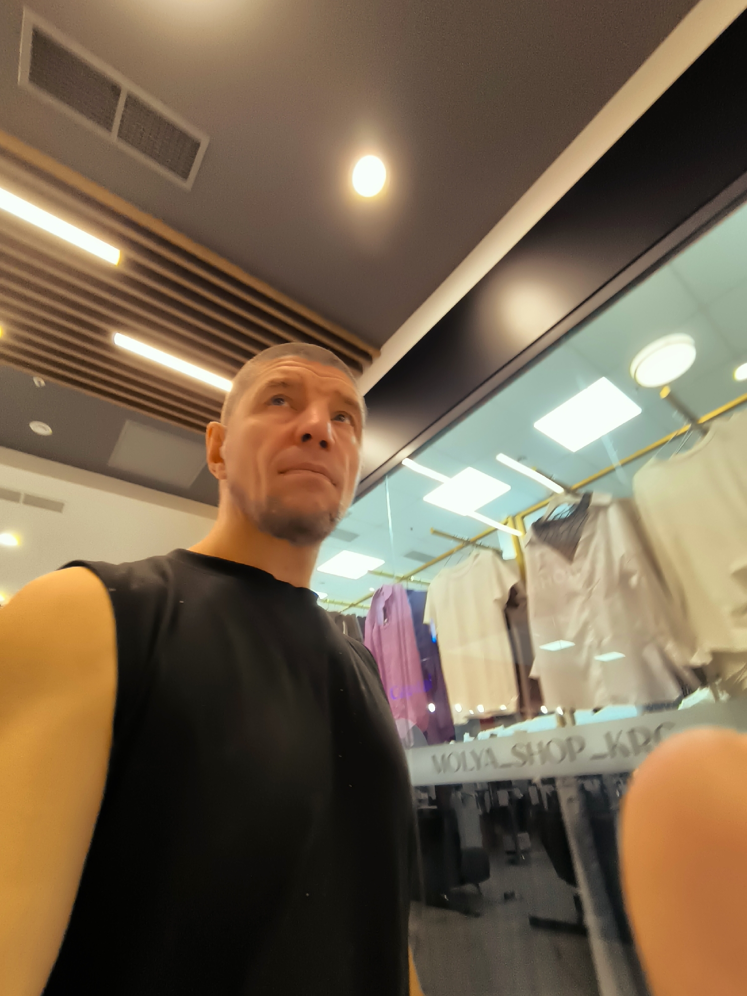 The image captures a man in a black sleeveless shirt looking upwards in what appears to be a clothing store. The store has modern lighting with prominent strip lights and ceiling panels. Behind the man, clothing items, predominantly white and light-colored tops, are displayed on racks behind a glass partition. A reflective sign on the glass reads "MOLYA_SHOP_KRG". The overall environment suggests an indoor retail setting. The image does not provide specific information or visual cues to confirm the location within Qarağandı city administration, Kazakhstan.