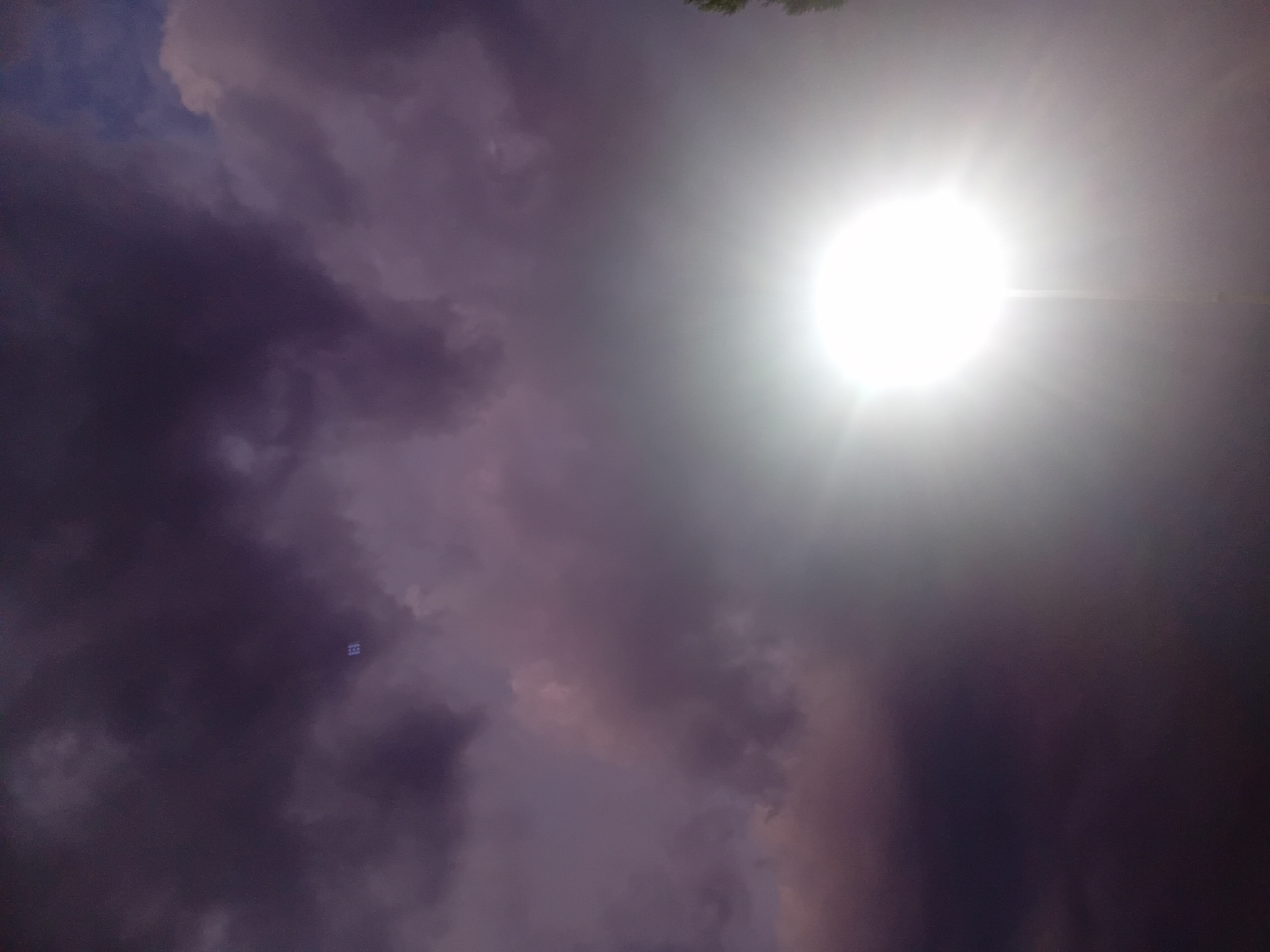 A bright, white light emanates from a streetlamp in the foreground of the image. The light is so intense that it creates a halo effect and obscures the lower portion of the streetlamp's pole. The sky is filled with dark, ominous clouds, tinged with purple and pink hues, suggesting twilight or stormy weather. In the upper left quadrant of the image, a small, square, blue object is visible amidst the clouds. A hint of dark foliage is visible on the far right edge of the image. The scene appears to be outdoors, potentially in an urban or suburban setting within the city of Onikişubat, Turkey, given the presence of a streetlamp. There are no people visible in the image.