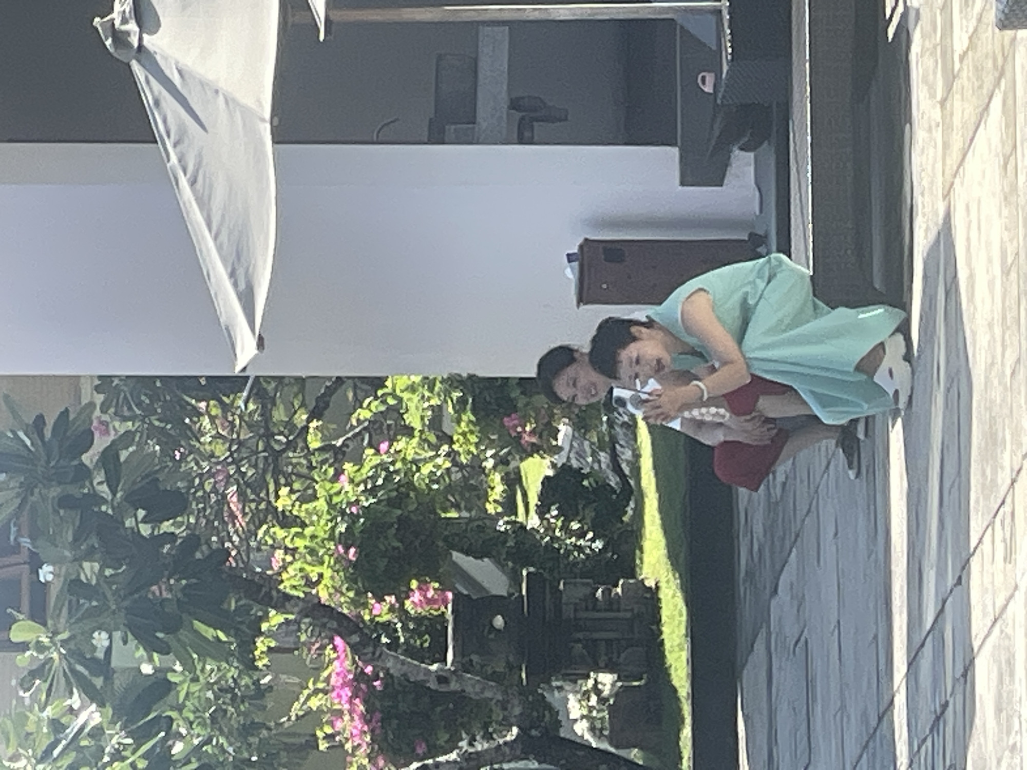 The image depicts two individuals in what appears to be an outdoor setting, possibly a resort or a residential area in Kuta Selatan, Indonesia. The scene is bathed in bright sunlight, suggesting it is daytime, and the weather seems clear.
In the foreground, there are lush green trees with vibrant pink flowers, indicating a tropical environment. A light-colored umbrella is partially visible in the upper left corner. The subjects are a person wearing a light blue dress and another person partially obscured, wearing red. The person in the blue dress is kneeling, facing towards the other person and holding what appears to be a white string of beads or pearls. The other individual is also leaning in, seemingly engaged in an activity with the person in blue. Their postures suggest a moment of interaction, possibly a demonstration or a shared activity.
In the background, there is a building with clean, modern architecture, featuring white walls and darker accents. A dark wooden cabinet is visible near the building. The ground is paved with light-colored tiles, reflecting the sunlight. The overall ambiance suggests a relaxed and serene atmosphere, possibly during a vacation or a cultural event. No text is visible in the image.