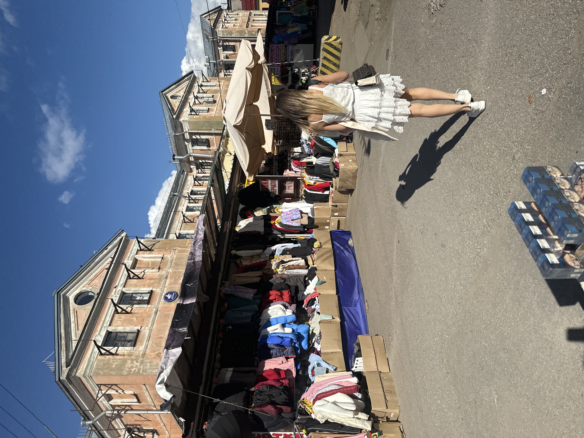 The image depicts an outdoor market scene in Saint Petersburg, Russia. In the foreground, a young woman with blonde hair walks away from the camera, wearing a white lace dress and white sneakers. She carries a black and beige shoulder bag. Her shadow is cast on the asphalt ground, which is littered with some debris. In the mid-ground, a clothing market stall is set up with racks of clothing, including jackets and shirts, displayed under a canopy. Cardboard boxes are stacked in front of the stall. Beyond the stall, a row of weathered, multi-story buildings with classical architectural elements stands under a bright blue sky with scattered clouds. The buildings have cream-colored facades and dark roofs. In the far background, more market stalls with awnings are visible. The overall impression is of a vibrant, sunlit market on a clear day.