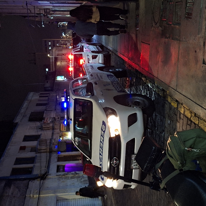 Night shot of a narrow street lined with parked white "SERENAZGO" vehicles with illuminated blue and red lights. People are walking on the sidewalk, and buildings are visible in the background. The foreground features part of a motorcycle with a green backpack. The street is wet, reflecting the vehicle lights.