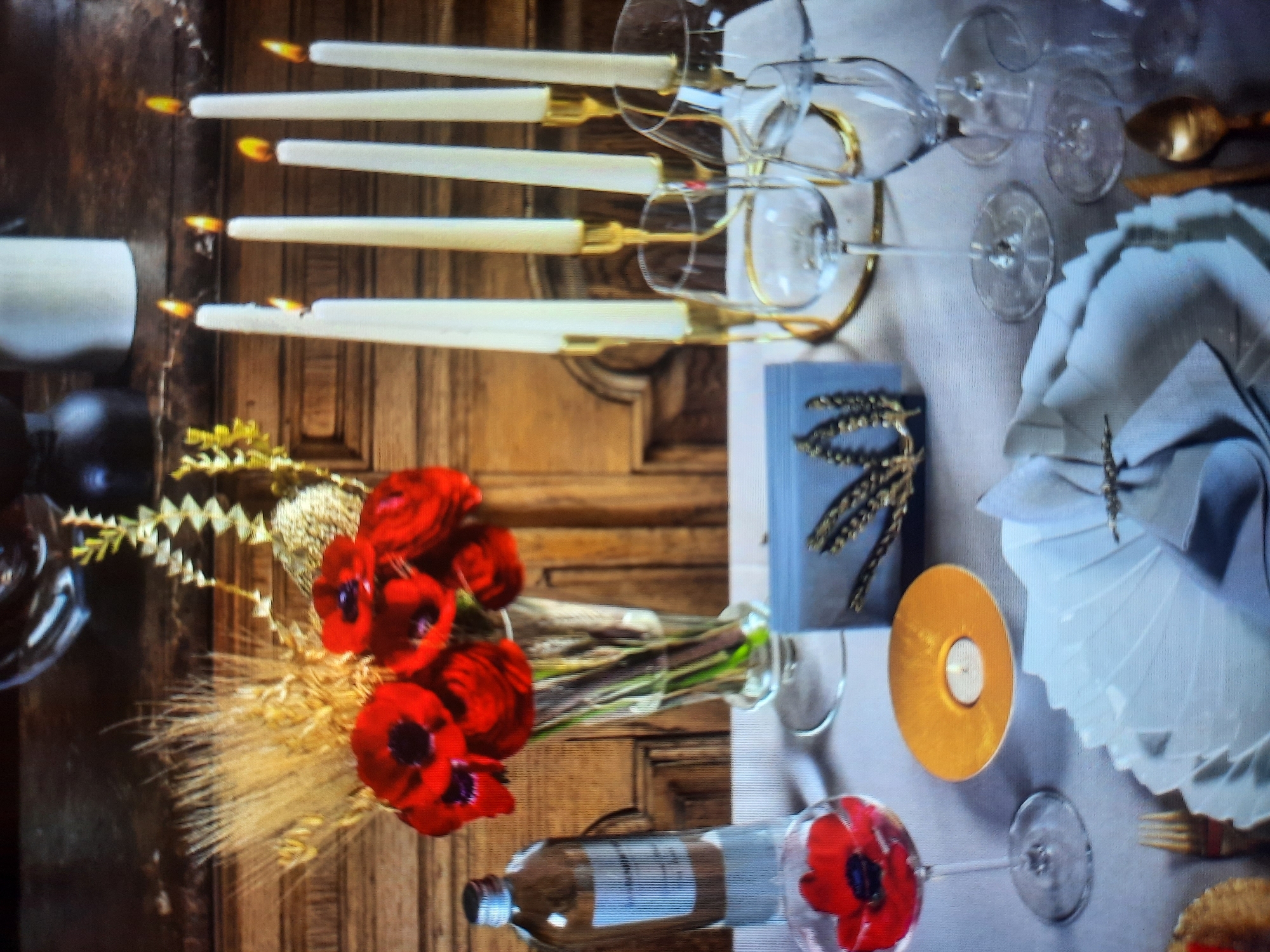 The image depicts a table setting with a focus on floral and candle elements, likely for an event or formal dining occasion. On the table, which is covered with a light-colored textured tablecloth, there is a clear glass vase containing a bouquet of red poppies and dried wheat stalks, appearing to be a centerpiece. To the left of the vase, a glass bottle with a label is visible.
In the foreground, a wine glass holds a single red poppy. Adjacent to this, a round, gold-colored holder contains a lit tealight candle. Further to the right, there are multiple tall, white taper candles with yellow flames, arranged in a gold candelabra that is partially obscured by wine glasses. These candles are placed within clear wine glasses, suggesting a decorative arrangement.
A grey card with a gold wheat stalk design is positioned on the table, likely a place card or menu holder. In the lower right quadrant, a white folded napkin, possibly made of linen, is secured with a decorative napkin ring. A golden spoon and a knife with a golden handle are also visible on the table. The background features wooden furniture, contributing to a warm and rustic ambiance.
This scene is not located in Rosenheim, Germany. The image depicts a tabletop setup that could be found in various dining establishments or homes internationally, with no specific indicators linking it to Rosenheim.