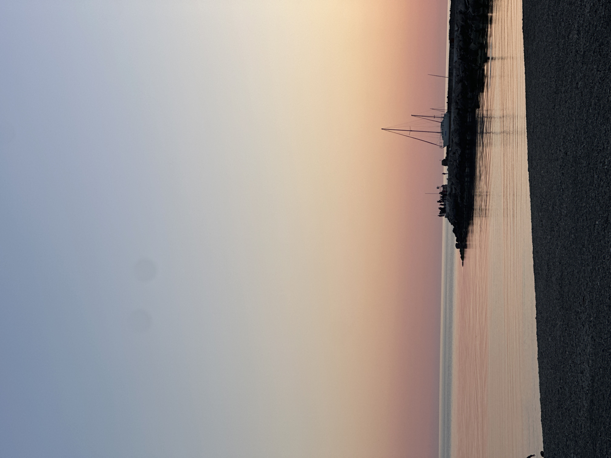 A landscape photograph, rotated vertically, depicts a shoreline at sunset. The sky transitions from a pale blue on the left to shades of orange and pink towards the right. The sea reflects these colors, creating a gradient effect. A dark rock breakwater extends into the sea, with several sailboats moored along its length. A dark grey beach is situated on the right side of the frame. Several figures can be discerned along the breakwater. The image appears to be taken in Novomikhaylovsky, Russia, judging by the breakwater and boats along the coast.