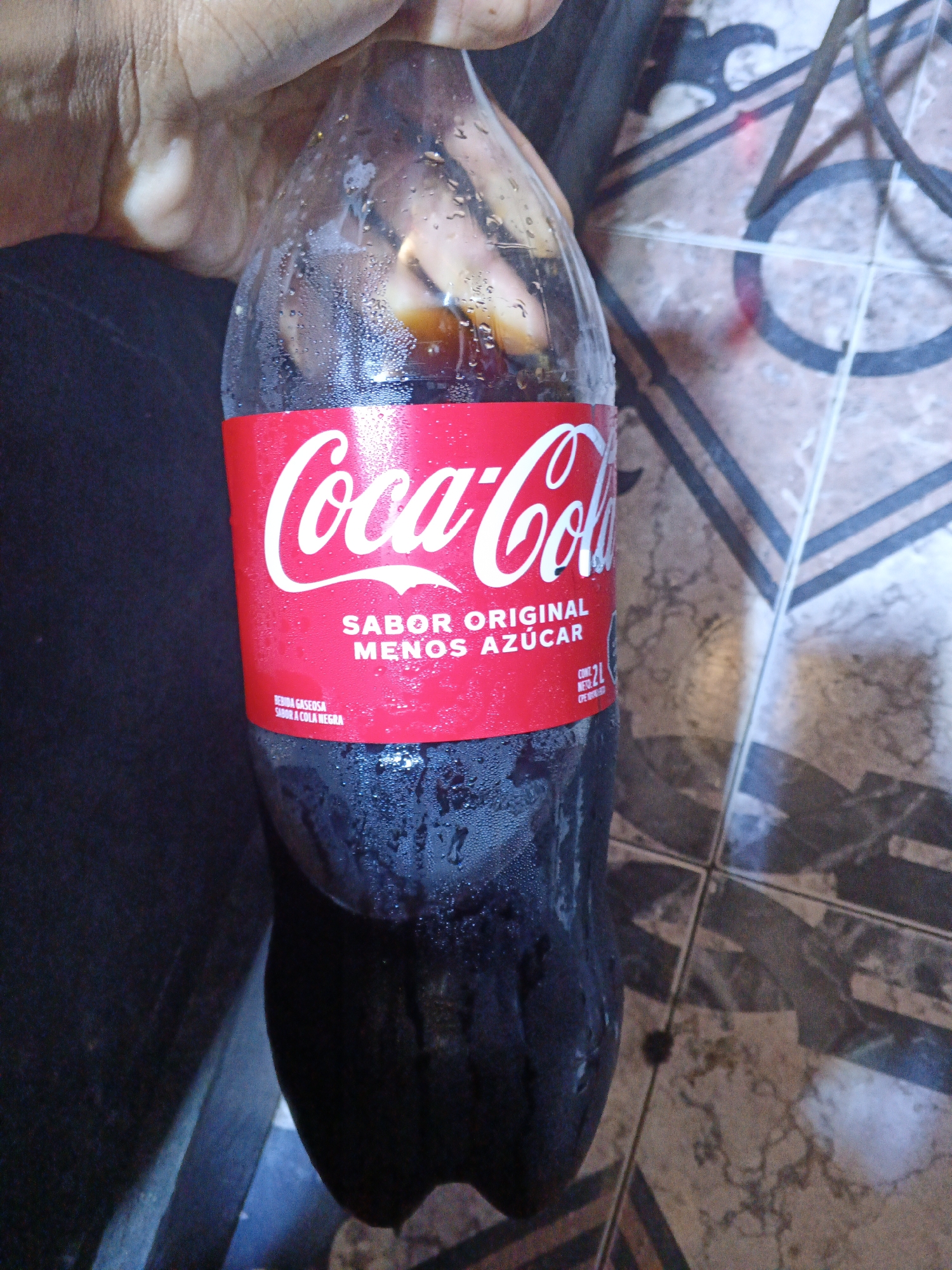 A person is holding a 2-liter bottle of Coca-Cola. The label indicates "Sabor Original Menos Azúcar" (Original Flavor Less Sugar) and "Bebida Gaseosa Sabor a Cola Negra" (Carbonated Drink Cola Black). The bottle is condensation-covered, suggesting it is cold. The background features a tiled floor and part of a metallic chair. The location is not specified, but it is a tiled indoor area. Altagracia, Venezuela, is not specified in the photo.