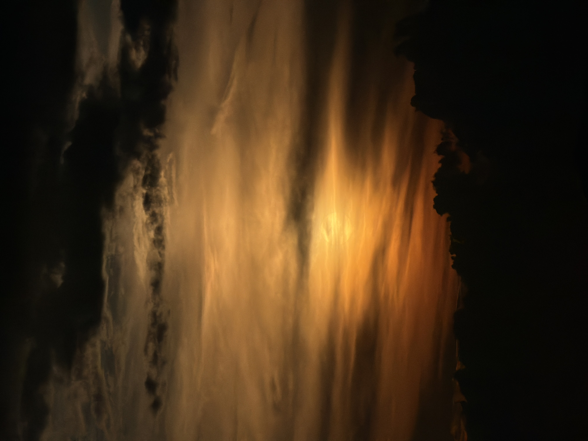 This is a photograph of a sunset over the city of Kuk, Montenegro. The sky is filled with dramatic clouds in shades of orange, yellow, and dark gray. The sun is a bright, glowing orb partially obscured by clouds, casting a warm light across the scene. The lower portion of the image is dominated by the dark silhouettes of clouds or possibly the outlines of buildings and trees in the foreground, creating a stark contrast with the brightly lit sky. The overall atmosphere is one of a powerful and dynamic sunset.