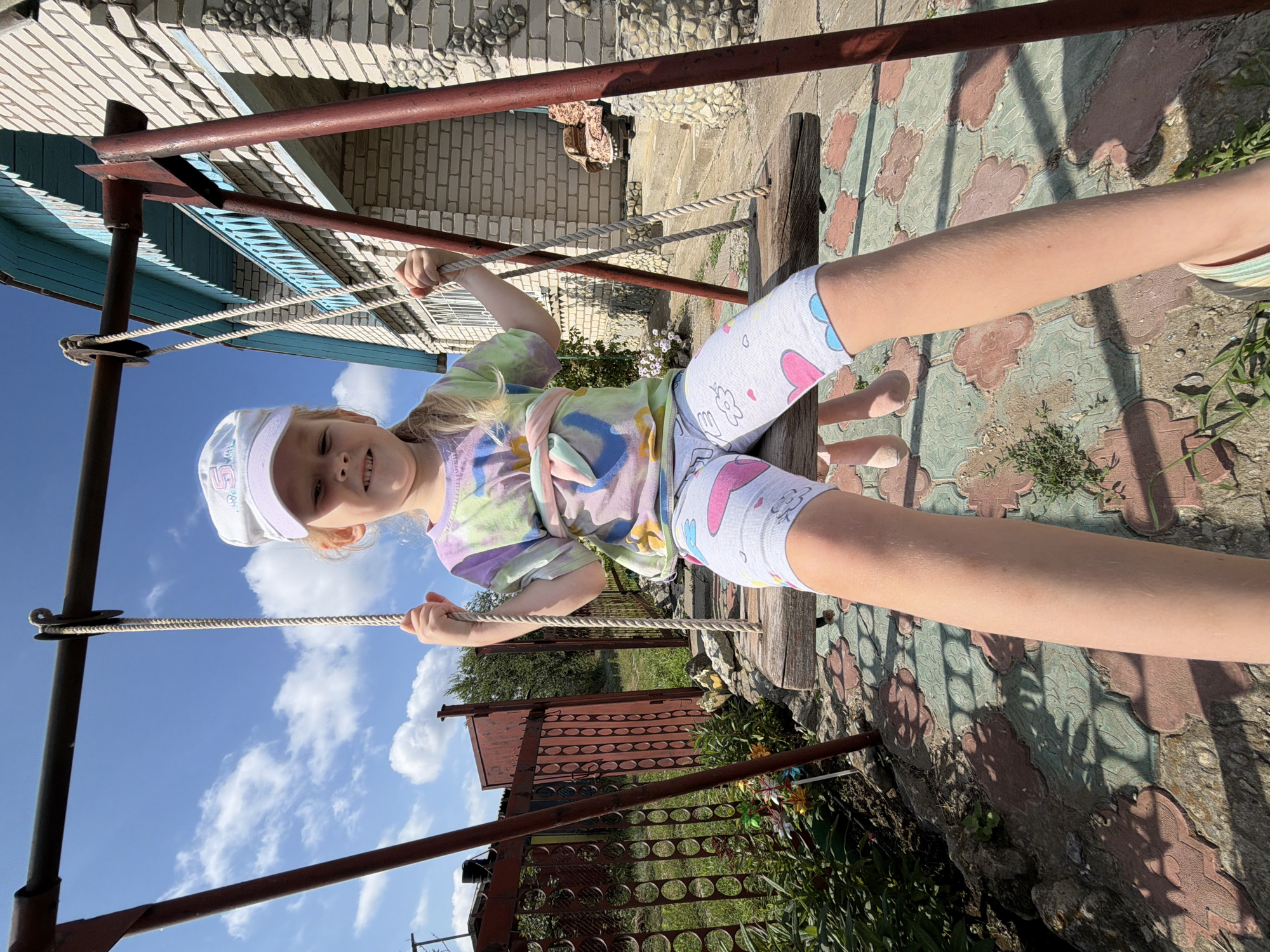 A young girl with blonde hair, wearing a tie-dye t-shirt, white shorts with cartoon prints, and a white baseball cap, is sitting on a wooden swing. She is smiling and looking towards the camera. Her arms are extended, holding onto the ropes of the swing. The swing is suspended from a metal frame.
The scene appears to be an outdoor setting, possibly a backyard in Kamyshin, Russia. The ground is paved with decorative tiles. In the background, there is a brick building and a fence. The weather is sunny with a blue sky and scattered clouds, suggesting it is daytime. The overall mood is cheerful and relaxed. There is no visible text in the image.