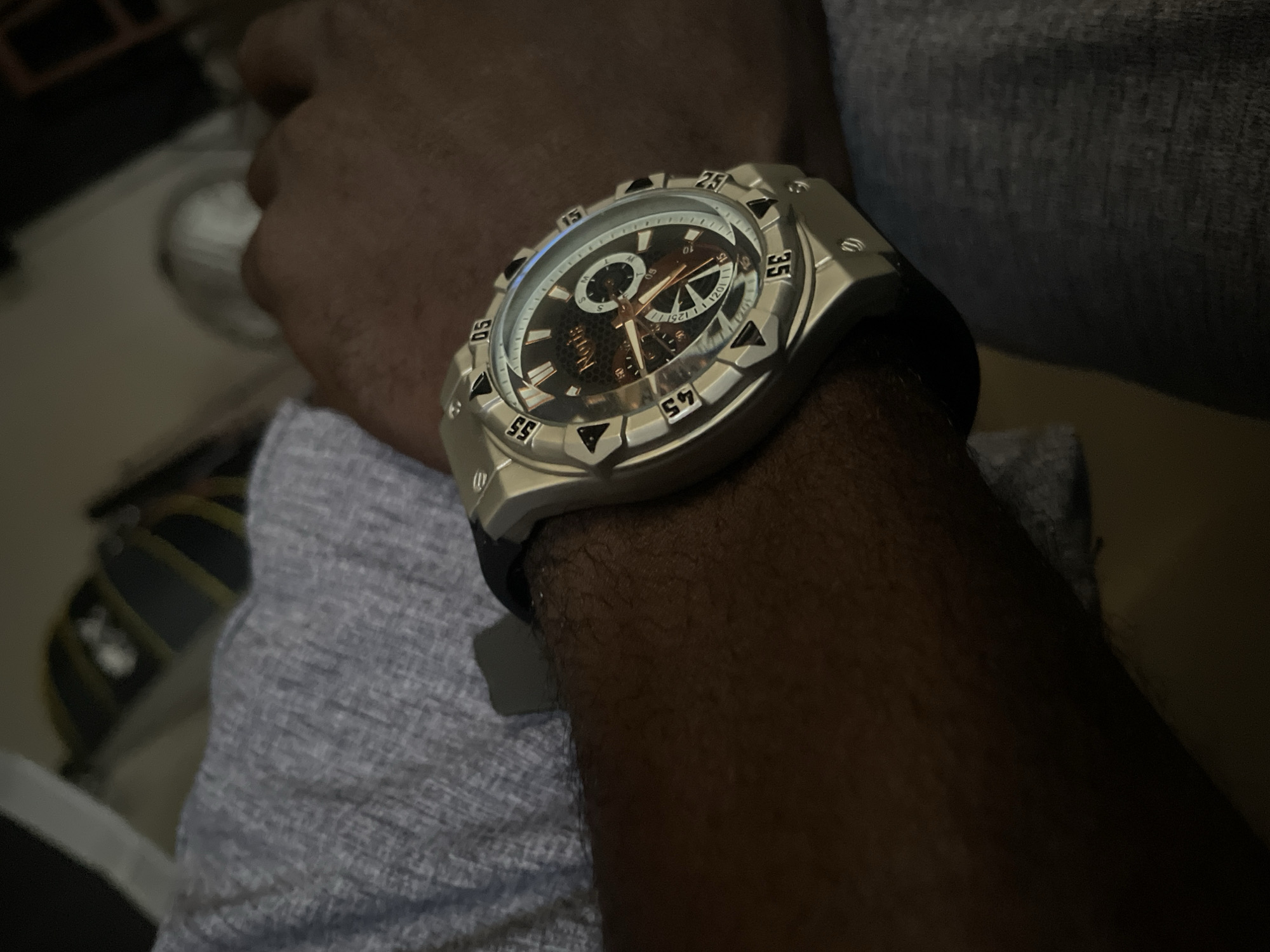 An extreme close-up shot shows a man's wrist and forearm. The man is wearing a silver-toned watch with a dark band. The watch face is dark with silver markings and hands. The man is wearing a grey t-shirt. The background is out of focus, but appears to be indoors, possibly in a living room or office. The image does not provide any information about the city of Durimi, Nigeria.
