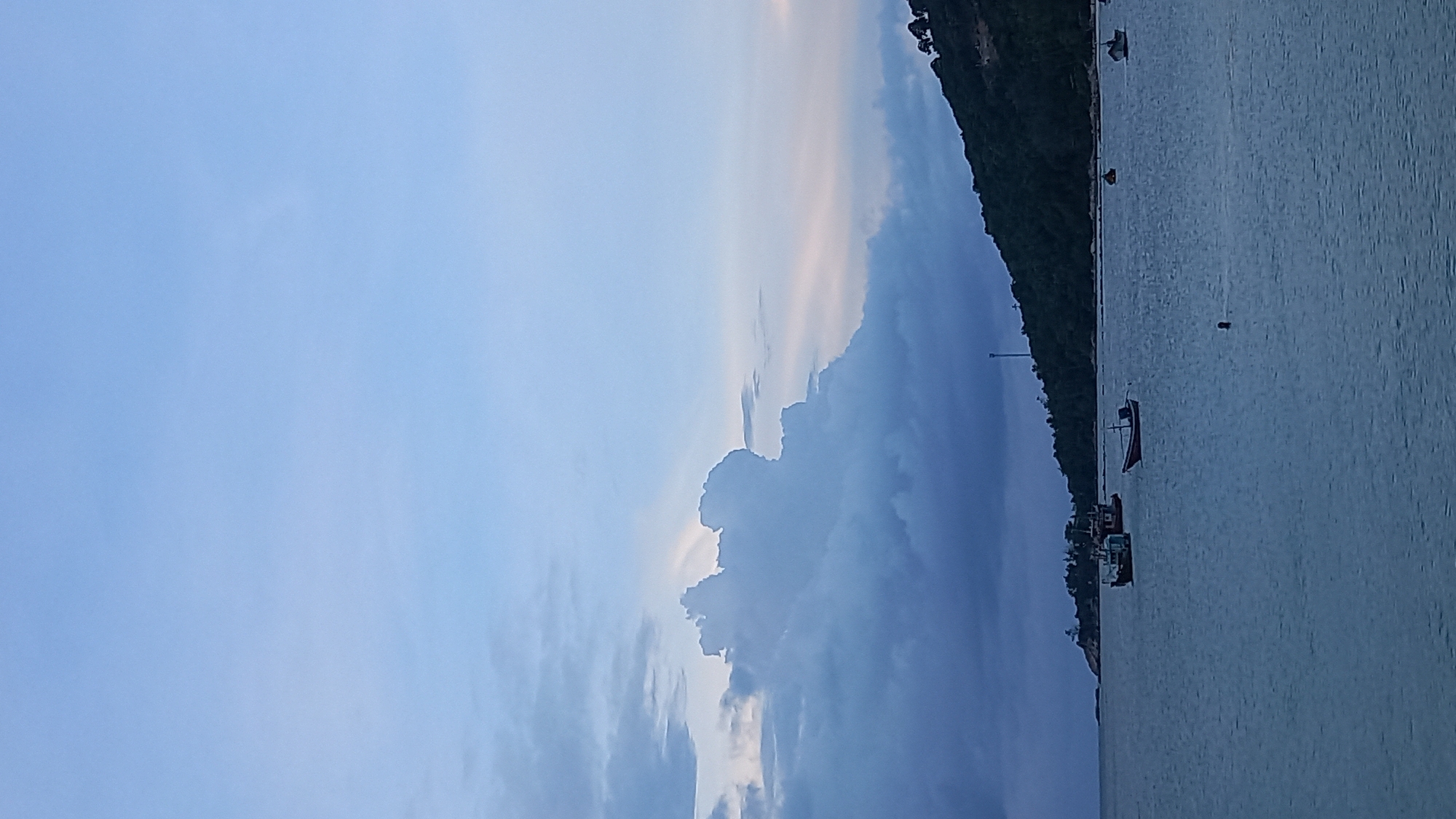 A wide-angle, vertical photograph captures a serene seascape at dusk in Ko Pha-ngan District, Thailand. The sky is a soft gradient of pale blue transitioning to a gentle pink near the horizon, with large, dramatic grey clouds dominating the left side of the frame. The sea is a calm, deep blue, reflecting the muted colors of the sky. Several small fishing boats are dotted across the water; three are in the middle distance, two clustered together, and one farther out to the right. A solitary, darker boat with a red trim is closer to the foreground. In the distance, a densely forested, dark green hill forms the coastline, its silhouette meeting the horizon. A tall, slender tower is visible on the hillside, with a few trees visible at its peak. The overall impression is one of peaceful twilight on the coast.