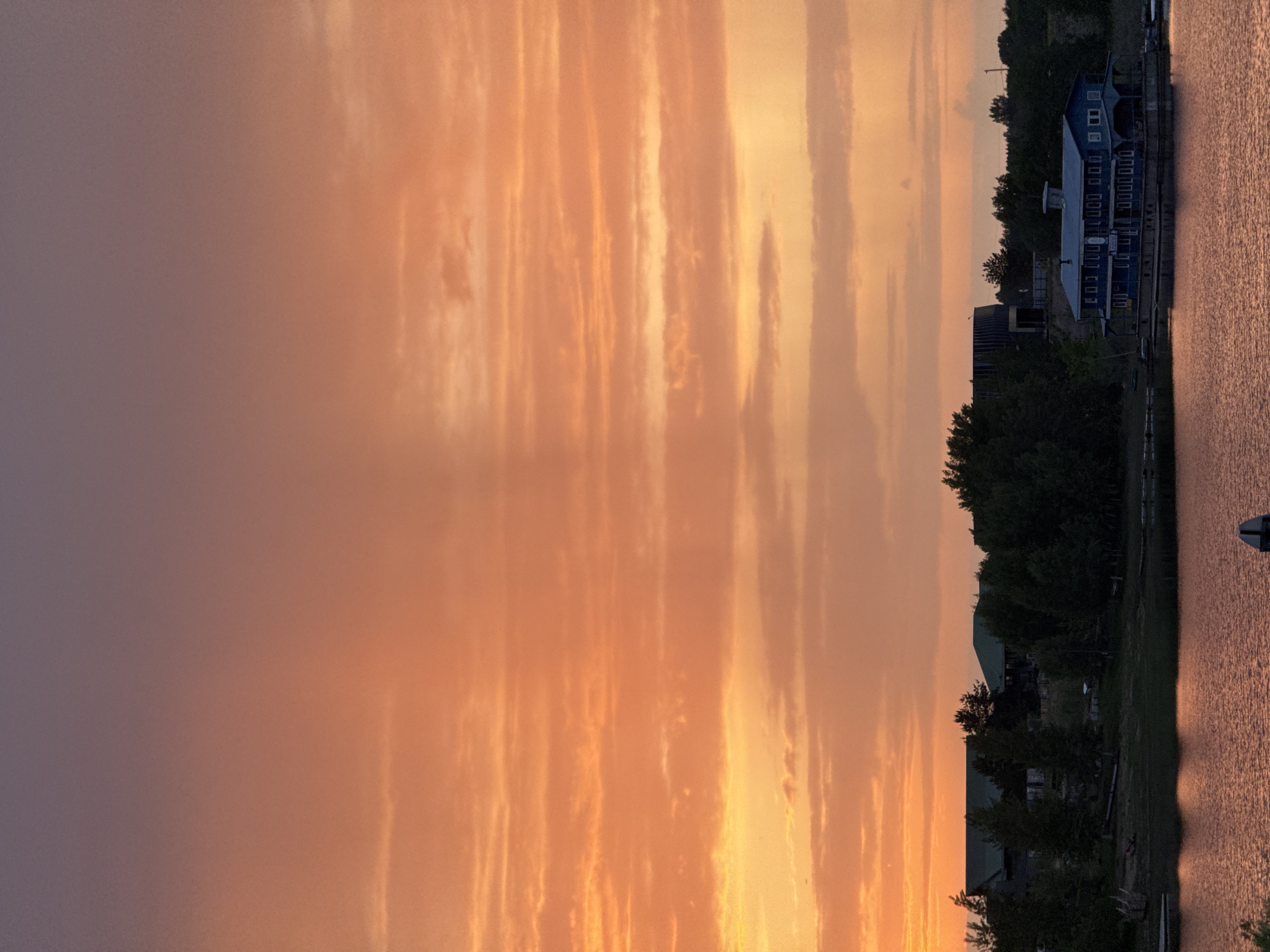 The image captures a serene sunset over a body of water, likely a lake or river. The sky is filled with a gradient of colors, transitioning from a deep purple or gray on the left to a bright orange and yellow on the right. There are vertical streaks of clouds that create a striated pattern across the sky. On the right side of the image, there is a shoreline with trees and buildings, including a large blue building with multiple windows that appears to be some kind of resort. The trees are dark, providing a contrast to the vibrant sky. The water reflects the colors of the sunset, with the surface appearing calm. The location is Nizhny Novgorod, Russia.