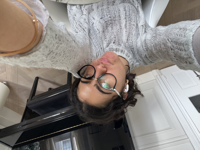 The image displays a female individual taking a selfie indoors. She possesses dark, curly hair, wears round black-rimmed glasses, and has a white earbud inserted in her left ear, along with a small gold hoop earring. Her facial expression is characterized by pursed lips. She is dressed in a light grey, intricately patterned, high-neck knitted sweater. A light brown strap, consistent with a bag or accessory, is visible across her upper left arm. The background elements indicate a residential interior in Paris, France, identifiable by characteristic white Haussmannian-style paneled walls and a light wood herringbone parquet floor. A black grand piano with a glossy finish and a gold accent trim is prominently positioned in the upper right, accompanied by a matching black padded piano bench. A portion of a white, conical floor object is visible on the far right. A white paneled door is present in the background on the left.