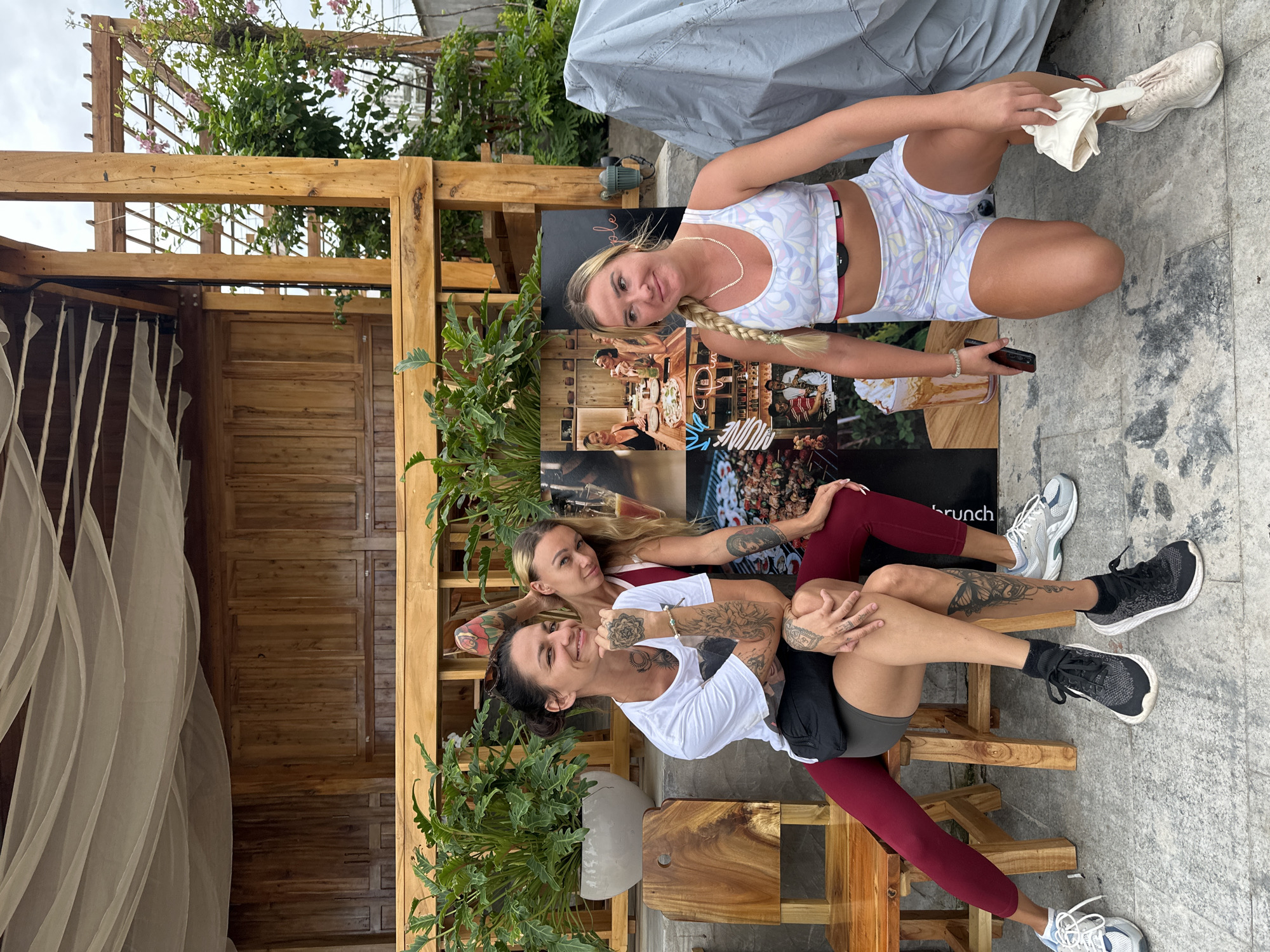 Three women are posing in front of a wooden structure with a canopy. The woman on the left is sitting on a stool, wearing a white t-shirt and dark shorts, with many tattoos visible on her arms and legs. The woman in the middle is leaning against the woman on the left, wearing a red top and dark leggings, also with visible tattoos. The woman on the right is kneeling, wearing a white sports bra and patterned shorts, with a heart rate monitor on her chest. They are all smiling and looking at the camera. Behind them is a large poster displaying various images, including a dining scene and food. In the background, there are plants and a wooden pergola. The ground appears to be paved with stone tiles. The overall scene suggests an outdoor gathering or photoshoot. The location is identified as Phu Thuy Ward, Vietnam.