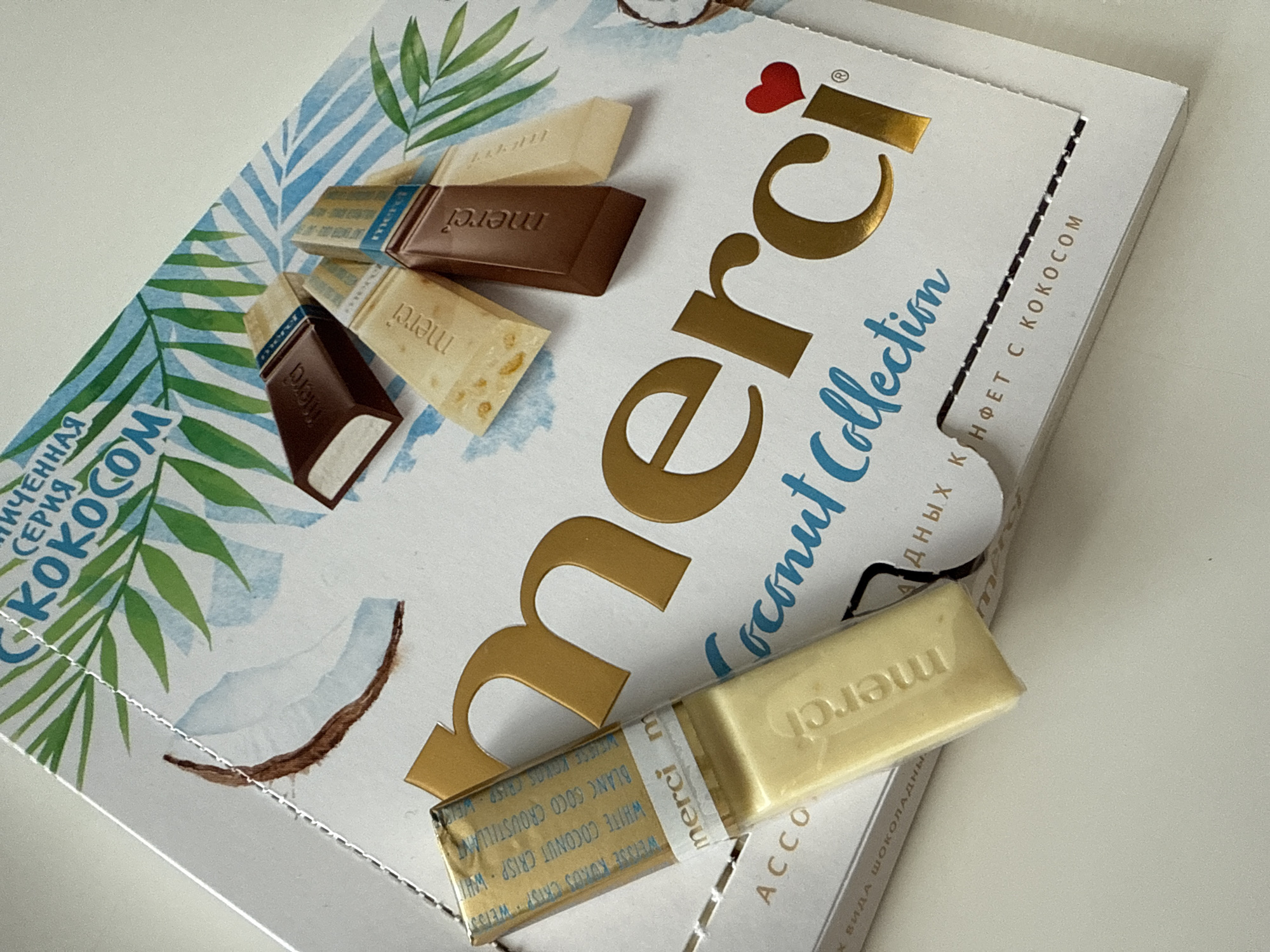 The image displays a close-up view of a Merci chocolate box, likely taken indoors on a light-colored surface in Lobanovo, Russia. The white cardboard box features a vibrant tropical design with watercolor-style green palm leaves and blue patterned stripes. An illustration of a sliced coconut is also visible on the packaging.
The prominent "merci" brand logo is rendered in metallic gold foil, accompanied by a small red heart and a registered trademark symbol. Below it, the phrase "Coconut Collection" is written in a cursive blue font. Several individual Merci chocolate pieces are arranged on and around the box. These include unwrapped rectangular chocolate bars, some in a darker milk chocolate color and others in a lighter, cream or white chocolate shade. The word "merci" is embossed on the surface of the unwrapped chocolates. One light-colored chocolate bar is partially encased in its gold-foiled wrapper.
Visible text on the box includes a partial Russian phrase "ЛИЧЕННАЯ СЕРИЯ КОКОСОМ" (part of "ОГРАНИЧЕННАЯ СЕРИЯ С КОКОСОМ" - Limited Series with Coconut) on the left side. On the right side flap, "АДНЫХ КОНФЕТ С КОКОСОМ" (part of "ШОКОЛАДНЫХ КОНФЕТ С КОКОСОМ" - chocolate candies with coconut) and "ВИДА ШОКОЛАДНЫ" (part of "6 ВИДА ШОКОЛАДНЫХ КОНФЕТ" - 6 types of chocolate candies) are legible. The wrapper of the partially exposed chocolate piece shows text such as "WEISSE KOKOS CRISP WEISS", "BLANC COCO CROUSTILLANT", and "WHITE COCONUT CRISP WHI", indicating a white coconut crisp flavor. The scene is well-lit, highlighting the textures and colors of the packaging and chocolates.