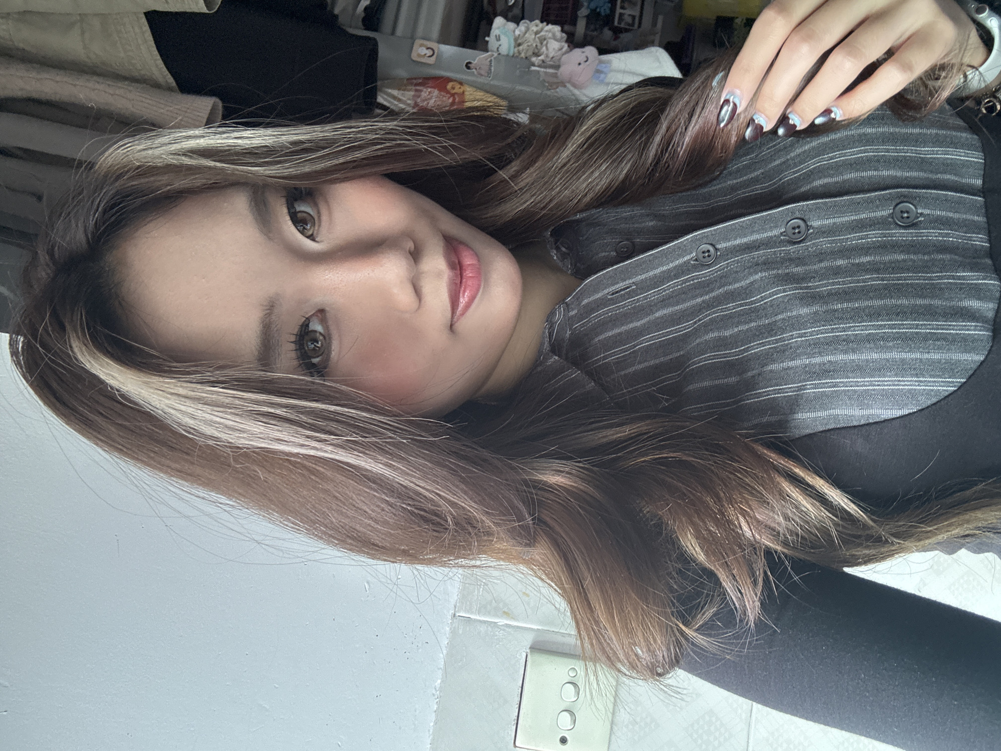 A woman with long brown hair with blonde highlights is facing the camera. She has brown eyes and pink lips. She is wearing a gray striped shirt with black buttons over a black long-sleeved top. Her left hand is visible, with a silver watch on the wrist and dark-colored nails with a silver and white design.
The background shows a cluttered room and a white wall with an electrical outlet in the lower right corner.
There is no information to indicate this picture was taken in the Nan'an District, China.