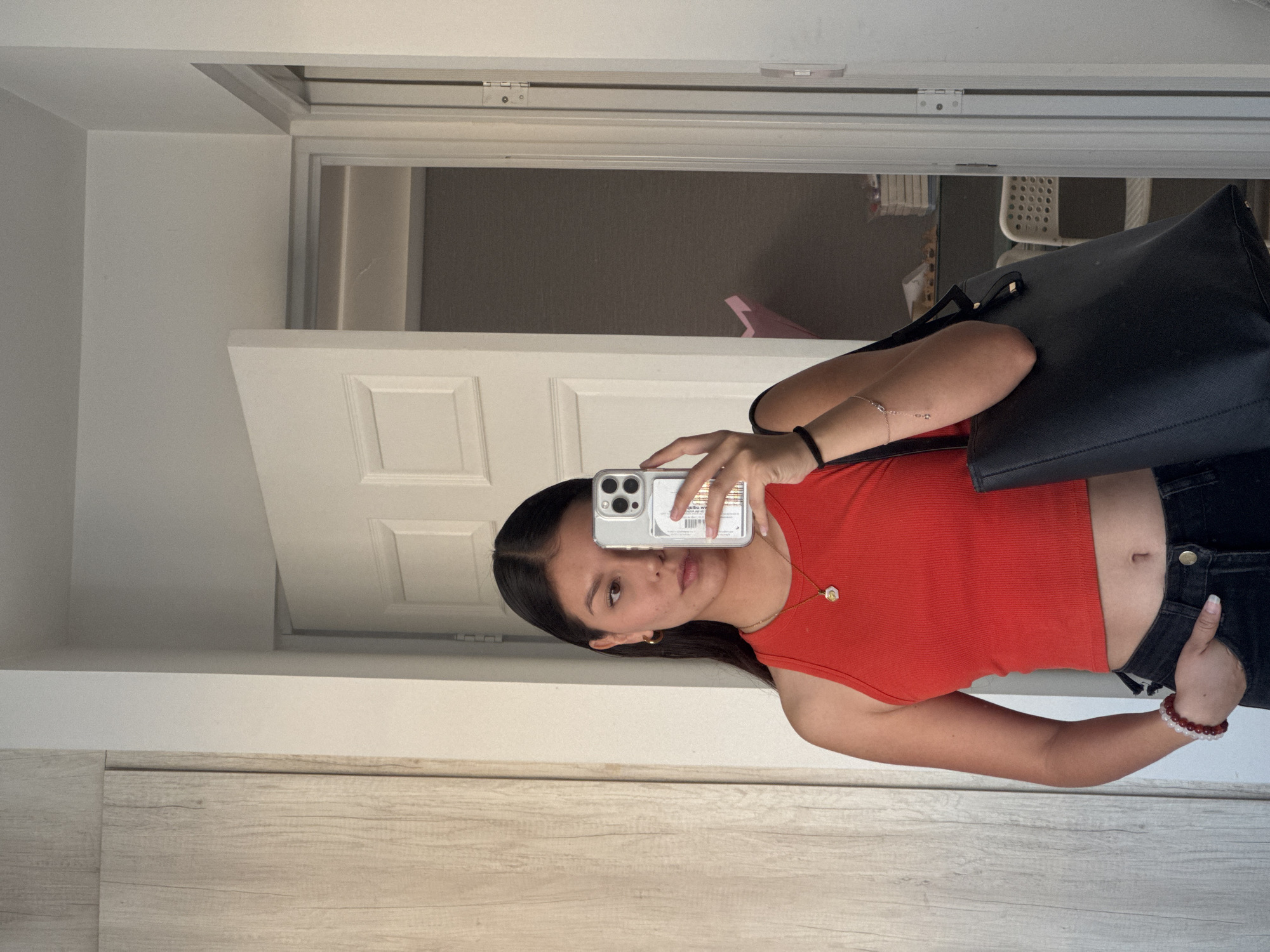 A young woman is depicted in a mirror selfie, standing indoors with a neutral expression as she looks towards the camera. She has long, dark hair and is wearing a ribbed, sleeveless red crop top paired with black jeans. Her left hand is casually placed in her jeans pocket. Accessories include a black elastic band on her right wrist, a red and white beaded bracelet on her left wrist, small hoop earrings, and a gold necklace with a small pendant. A black tote bag is carried over her right arm.
She holds a silver smartphone, likely an iPhone, in a clear case, which reflects her image. On the back of the phone case, reversed text is visible, reading "Malibu.w" with "California" underneath, suggesting a design or sticker.
The setting is an indoor space, characterized by white walls, a white paneled door, and a light wooden floor or baseboard. Through an open doorway reflected in the mirror, elements of another room are visible, including what appears to be a white plastic chair and some indistinct objects. The lighting suggests it is daytime. The provided location context is San Francisco Ocotlán (Ocotlán), Mexico.