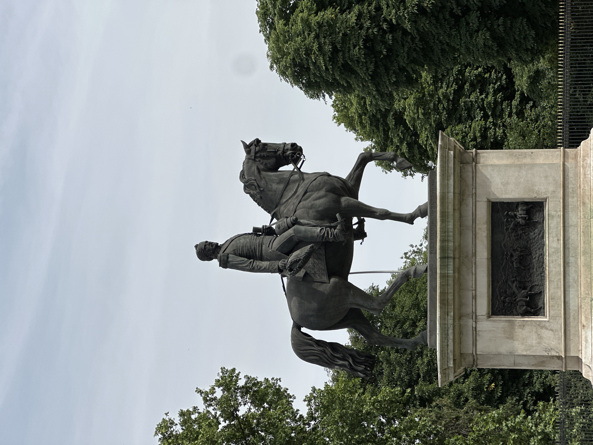 The image depicts the equestrian statue of King Charles III in Madrid, Spain. The bronze statue shows the monarch on horseback, with the horse rearing up on its hind legs. The statue is positioned on a large stone pedestal, which is decorated with bas-relief sculptures. The sky is partially visible in the background. The statue is surrounded by trees and a wrought iron fence.