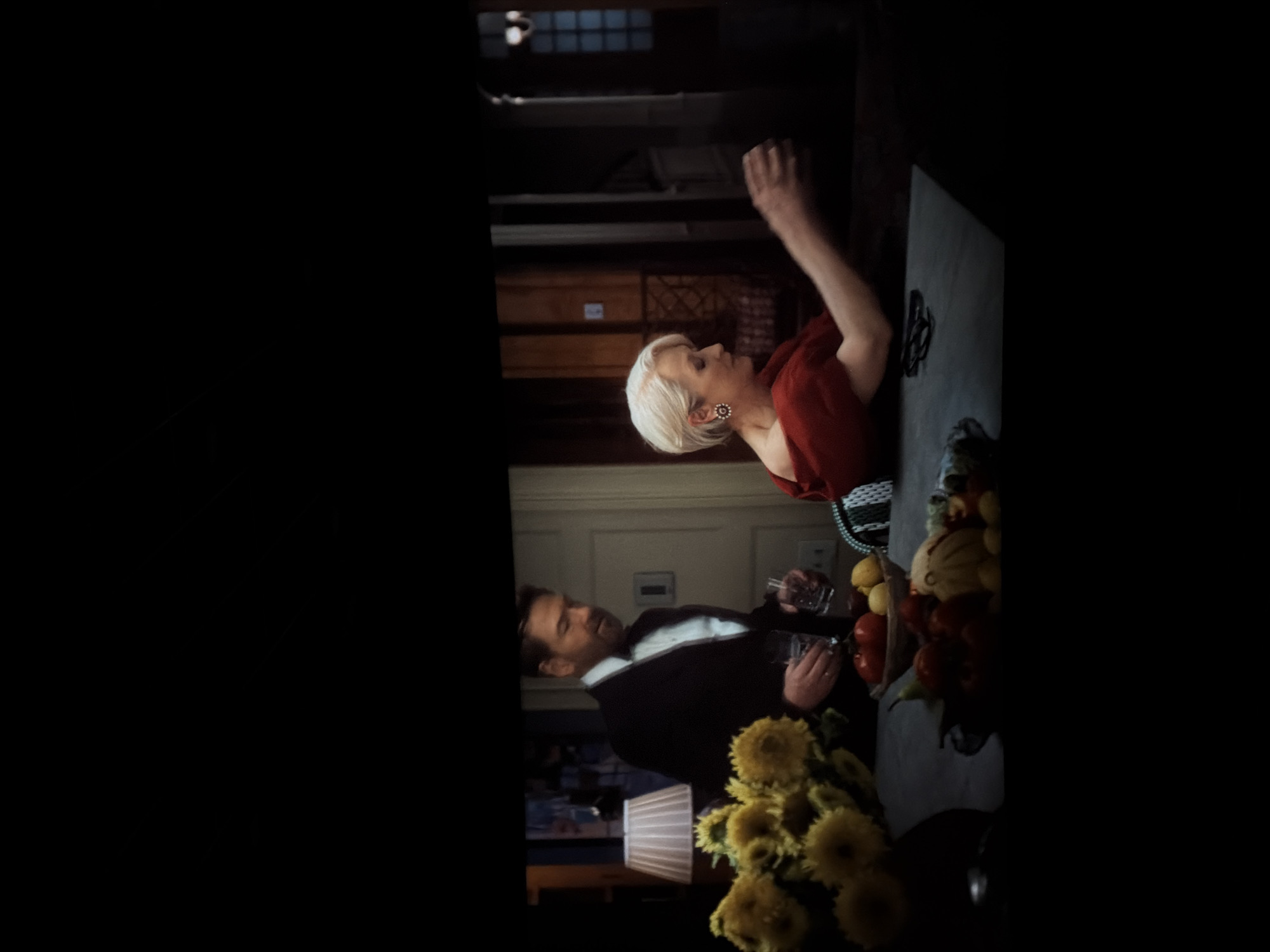 The image depicts a scene indoors. A man in a dark suit and white shirt is holding a glass. A woman with short, blonde hair and wearing a red top with earrings is standing next to him. They are near a table or counter laden with fruit, flowers, and decorative items. The background suggests an interior setting with doors, cabinets, and other home decor elements.
There is no evidence suggesting the location is in Yerevan, Armenia.