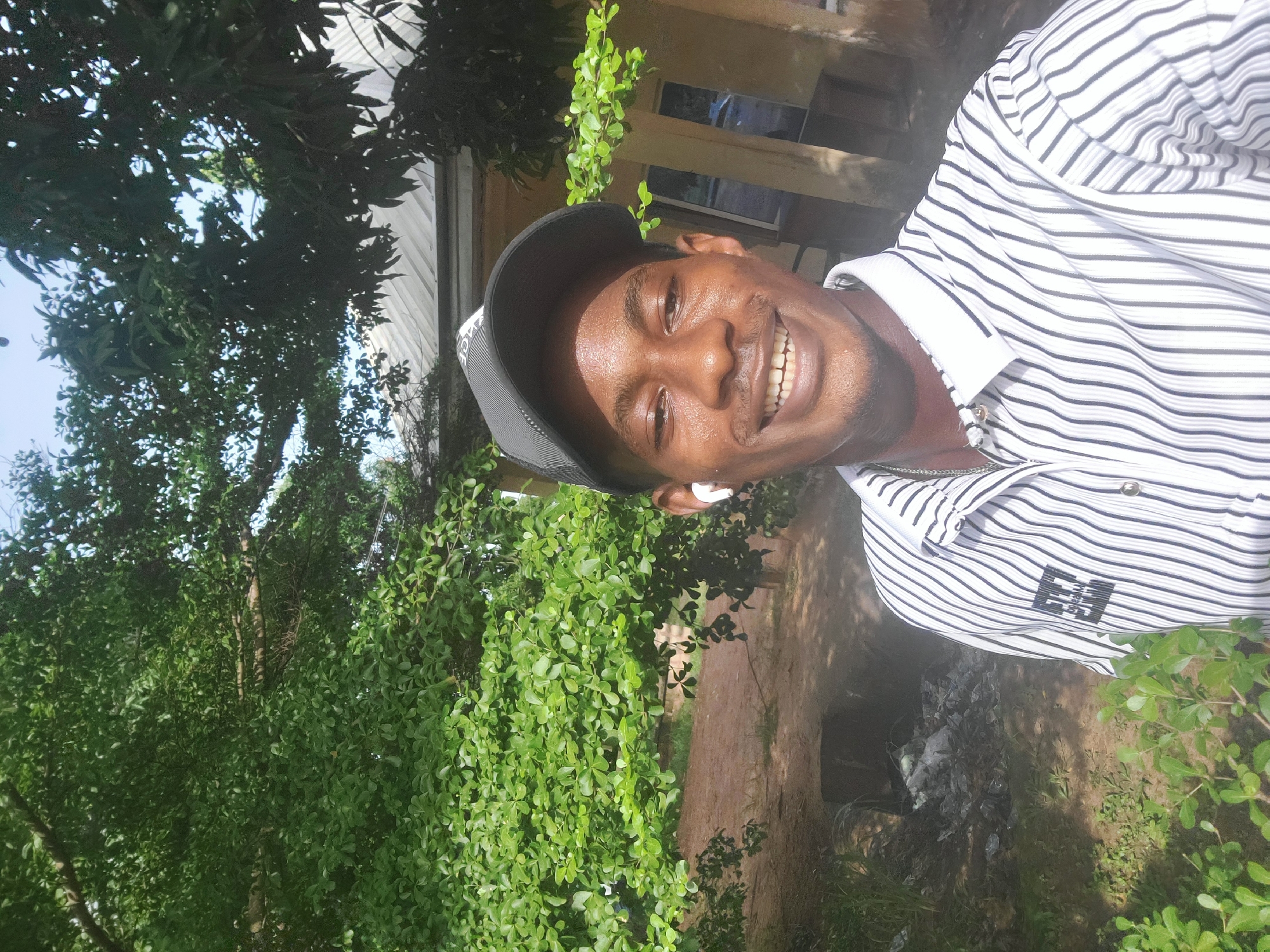 A man wearing a black baseball cap and a white and black striped collared shirt takes a selfie outdoors. He is smiling and has white earbuds in his ears. In the background, there are trees with green foliage, a building with a metal roof, and some scattered debris on the ground. The scene appears to be in Ukukwa, Nigeria.