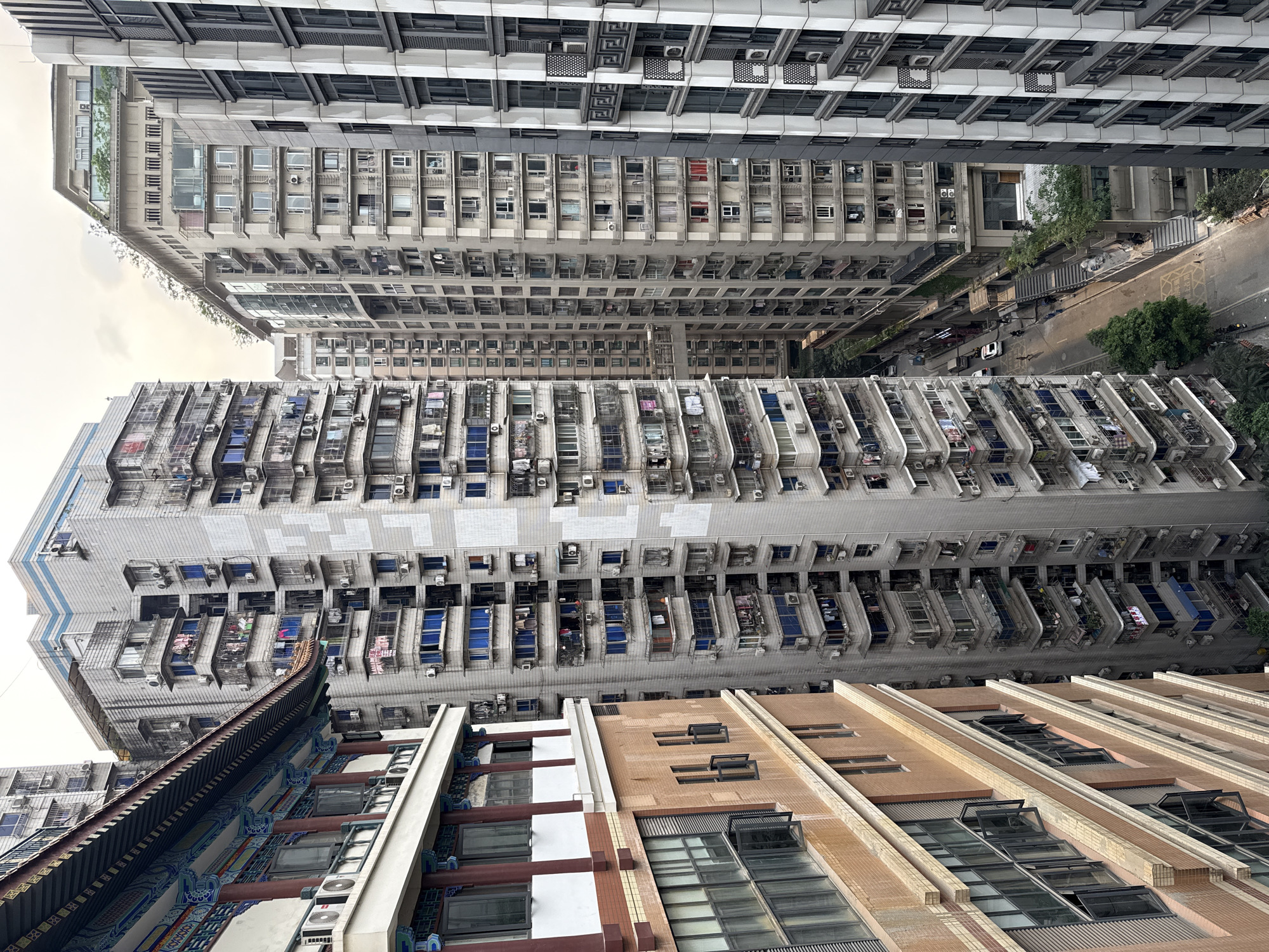 The image shows a high-angle view of densely packed residential buildings in Yuzhong District, China. The buildings vary in height and architectural style, ranging from older, tiled structures with traditional Chinese roof elements to modern high-rises with numerous air conditioning units. Clotheslines are visible on balconies, indicating domestic activity. A street with some vehicles and trees can be seen between the buildings. The overall scene depicts a densely populated urban environment.