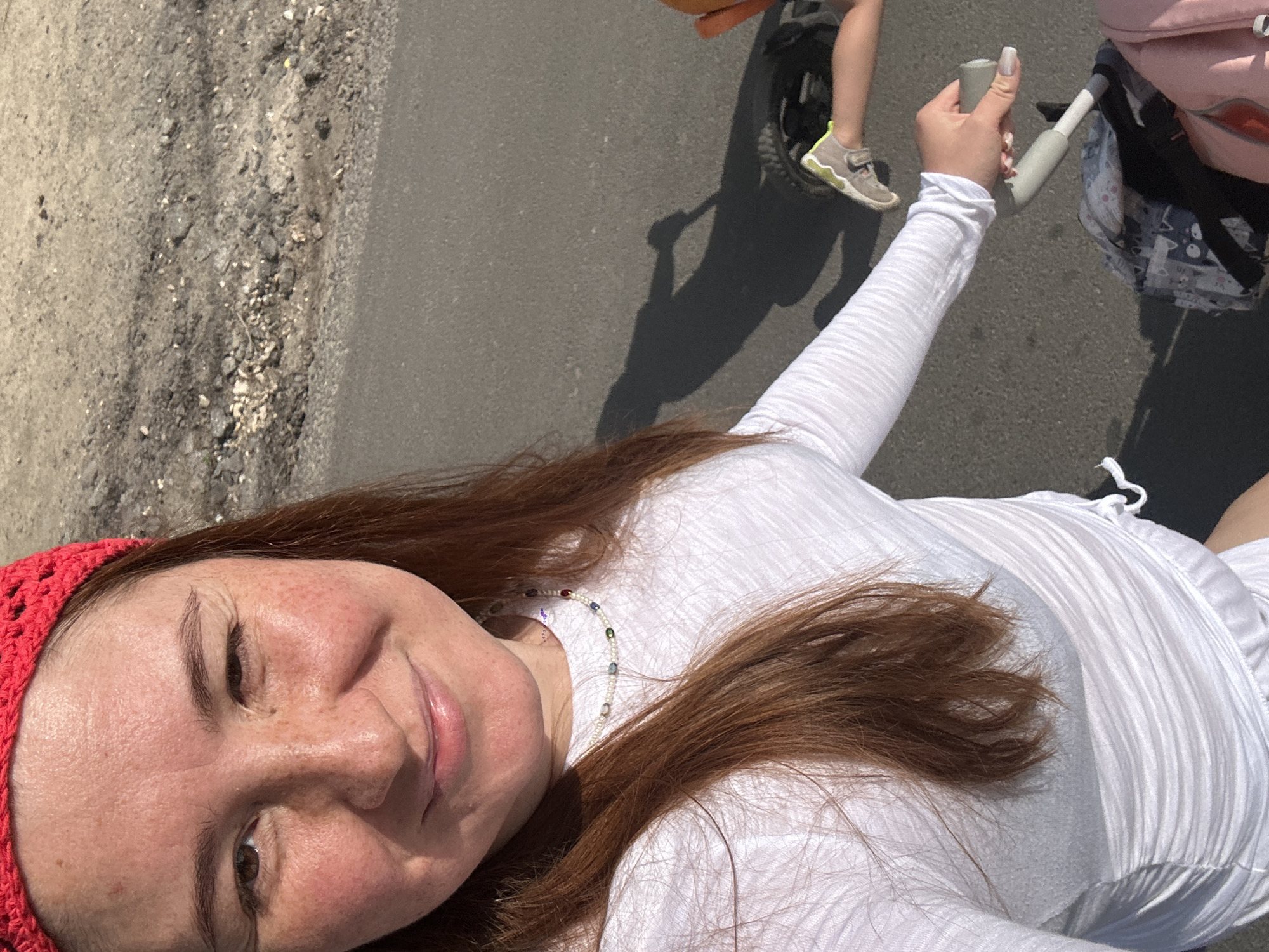 The image shows a woman with reddish-brown hair, wearing a red headband and a white long-sleeved shirt. She is outdoors, on an asphalt surface with gravel and dirt to the side. Her right hand is on the handle of a stroller, suggesting she is pushing it. A child's leg with a gray and yellow shoe is visible near the stroller's wheel. A shadow of the woman and the stroller is cast on the ground. The scene appears to be a sunny day. The image does not provide enough information to determine the location in the city of Nizhny Novgorod, Russia.