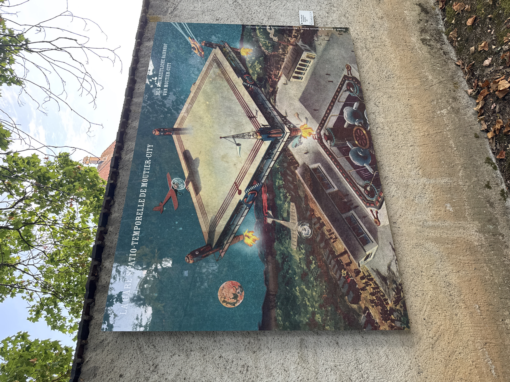 The image depicts a large, weathered mural on a concrete wall, likely outdoors. The mural is an illustration with a vintage, science-fiction aesthetic, featuring a complex, multi-layered structure resembling a train station or a futuristic city. The artwork has text in French and German. The French text on the left reads "LA GARE SPATIO-TEMPORELLE DE MOUTIER-CITY". The German text on the right reads "DER RAUMZEITLICHE BAHNHOF VON MOUTIER-CITY". This translates to "The Spatio-Temporal Station of Moutier-City" and "The Spatio-Temporal Station of Moutier-City" respectively, indicating the subject of the artwork.
The mural itself is detailed and colorful, despite some signs of age and weathering. It shows a central platform with a boxing ring-like structure, supported by elaborate mechanical legs that appear to be emitting flames. Above this platform, an orange airplane is depicted. In the background, there are stylized mountains and a reddish, cratered planet in the sky. The lower part of the mural shows buildings, what appear to be train tracks with a train, and more industrial-looking machinery. Figures, possibly people, are depicted in various parts of the mural, suggesting activity.
Attached to the wall to the right of the mural is a small white information plaque. The plaque is difficult to read in its entirety but contains some text that appears to be in French and possibly includes information about the artwork's creator or context. The title of the artwork itself refers to "Moutier-City," which is a fictional location.
The mural is mounted on a rough, textured concrete wall, painted a neutral, earthy tone. Above the wall, branches of green leafy trees are visible against a bright sky with some clouds. Some fallen brown leaves are scattered on the ground at the base of the wall.
Based on the textual reference to "Moutier-City" and the context of public art installations, this mural is likely located in or related to the region of Moutier, which is in the canton of Bern, Switzerland, and close to the canton of Jura, where Porrentruy is located. It's possible this artwork is part of a public art project in the vicinity of Moutier or a related town, and the inclusion of "Moutier-City" is a conceptual artistic choice rather than a literal geographical location. The overall impression is of an outdoor urban or semi-urban environment with a focus on historical or artistic display.