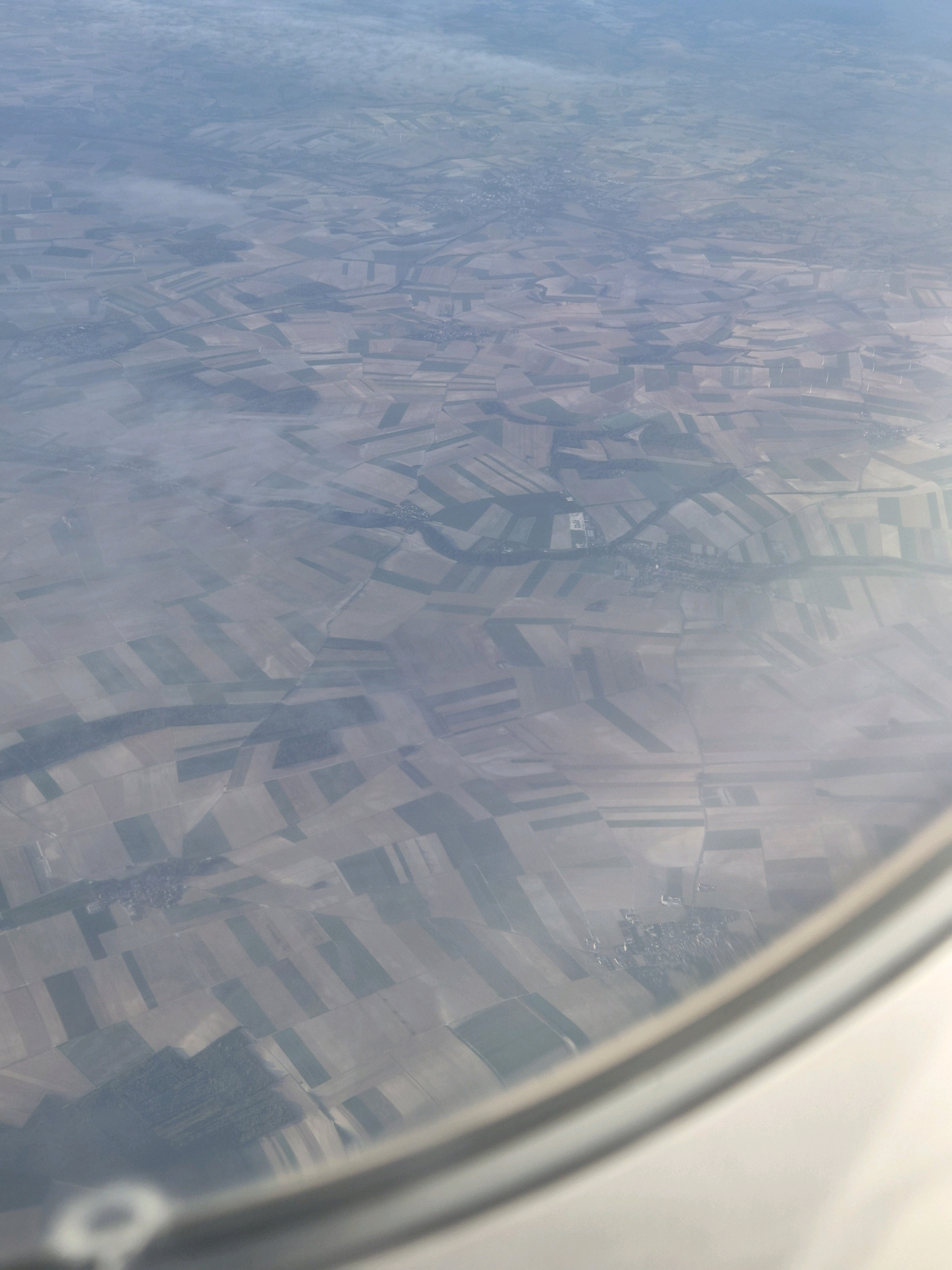 The image is an aerial view of a landscape taken from an airplane window, showing a patchwork of agricultural fields. The fields are varied in color and texture, indicating different crops or stages of cultivation. Some fields are rectangular, while others follow more irregular shapes. A small village, identified as Saint-Hilaire-le-Petit, France, is visible in the center of the image, with buildings clustered together. There are also some areas of trees and possibly wind turbines scattered throughout the landscape. The view is somewhat hazy, suggesting the presence of clouds or atmospheric conditions affecting visibility. The bottom right of the image shows a portion of the airplane's window frame, curved and light-colored.