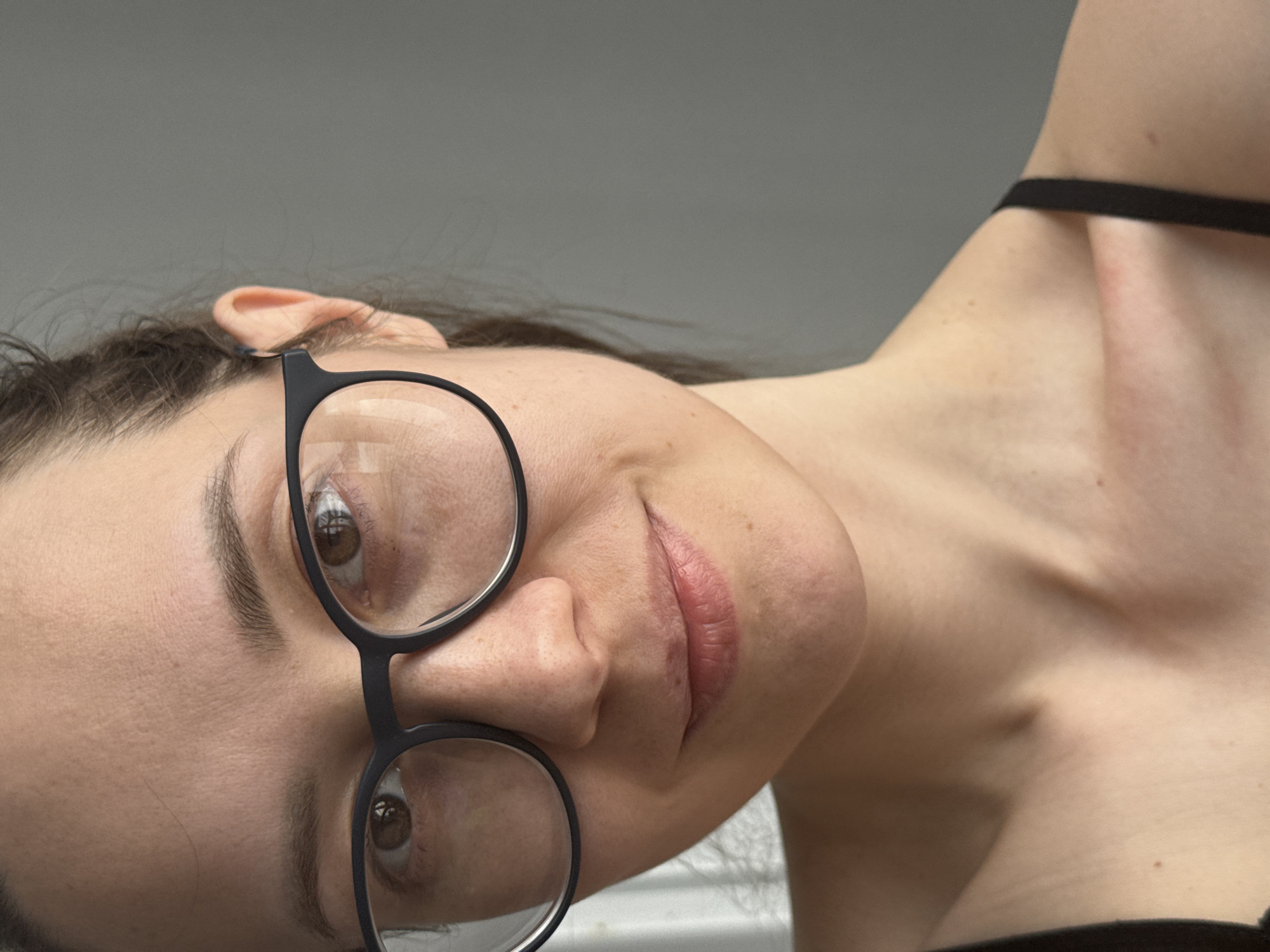 A close-up shot features a person's face, angled with their head tilted. The person wears black-rimmed glasses. A portion of the person's neck and shoulder area is visible, where a thin black strap rests. The background is a blurred, pale grey. There is no indication of a location in the city of Chaikovsky, Russia.