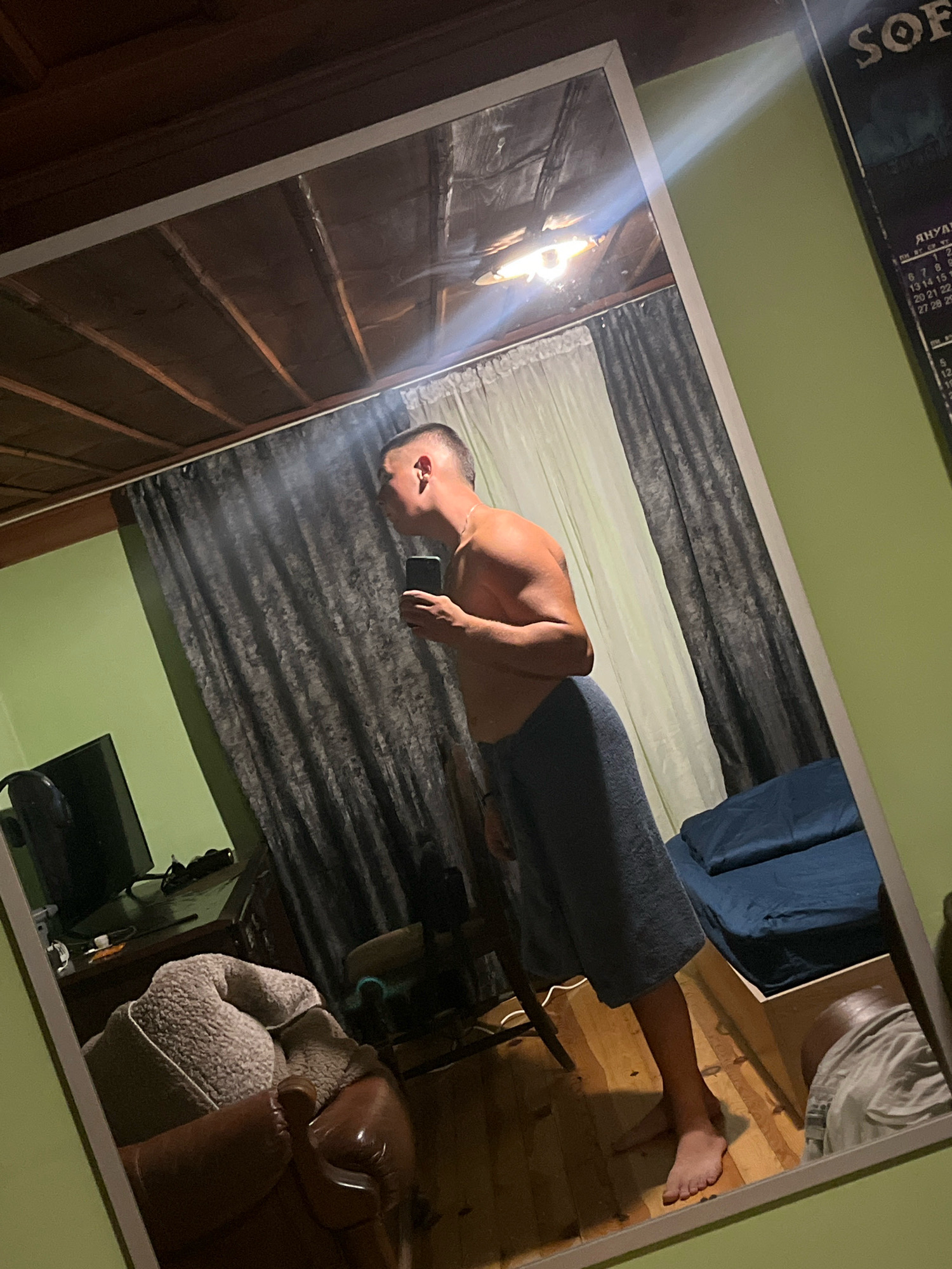 A full-body mirror reflects a young man standing in a room. He is shirtless, wearing a blue towel wrapped around his waist, and holding a smartphone in his right hand. The mirror is positioned at an angle, capturing a portion of the room's interior. The room appears to be a bedroom, with a wooden ceiling, green walls, and dark curtains. In the reflection, a bed with blue bedding is visible, along with a TV set and furniture. The floor is made of wood. There is a calendar on the wall to the right of the mirror, partially visible. The man is looking into the mirror, and his expression is neutral. The lighting in the room is dim, with a light source reflecting off the ceiling. The overall impression is of a casual, personal moment captured in a private space.