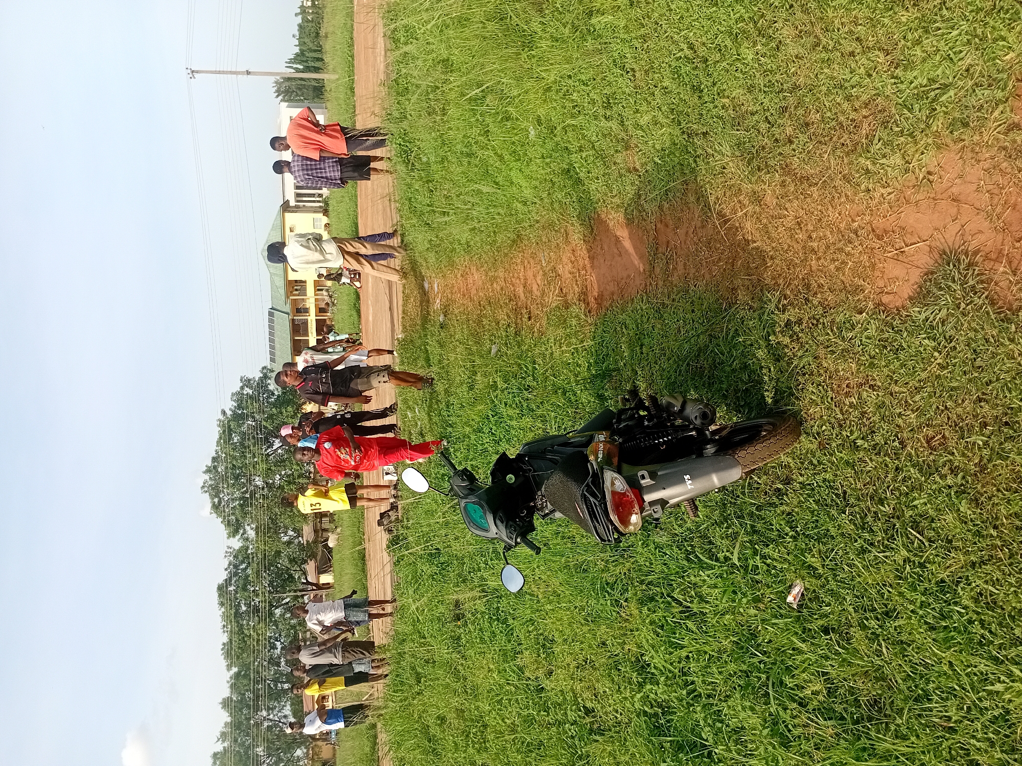 The image shows a horizontal view of a grassy field next to a dirt path in Ogbogbo, Nigeria. A black motorcycle lies on its side in the grass. A group of people are standing near the path. Some of them are wearing sports jerseys in red and yellow, while others are in casual clothes. A building and power lines are visible in the background.
