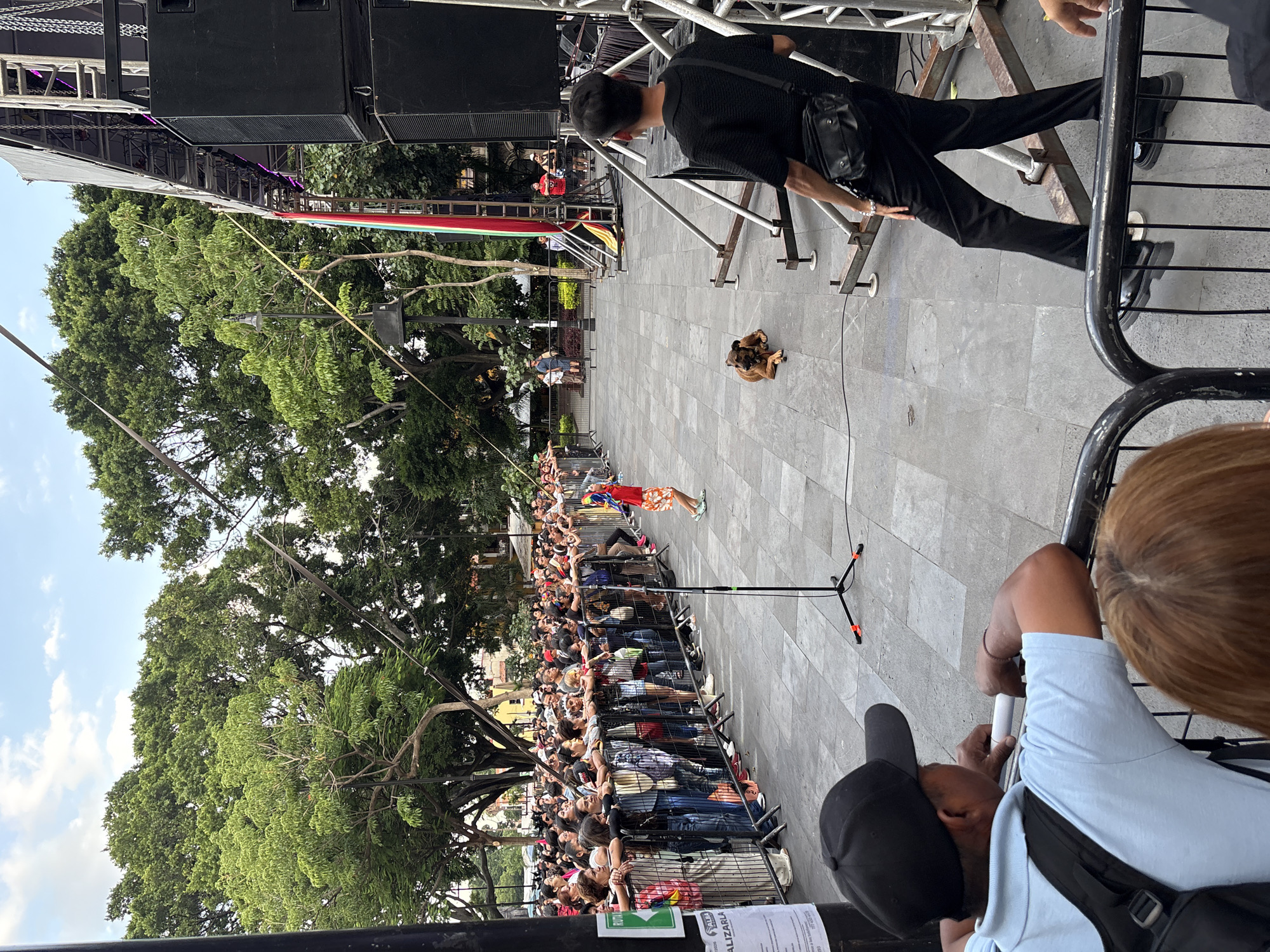 The image depicts an outdoor event in Cuernavaca, Mexico, likely a public gathering or performance. A crowd of people is visible behind a metal barrier, watching a performance or presentation. In the foreground, a man wearing a light blue shirt and a black baseball cap leans on the barrier, looking towards the event. To his right, a woman with reddish-brown hair is also partially visible.
Further into the scene, a young person in an orange and red outfit stands in the open space, facing away from the crowd and towards a stage structure. A brown dog is lying on the paved ground near the stage. The stage itself is large, with multiple speakers and lighting equipment, and is decorated with colorful banners, including red, green, and white, suggestive of the Mexican flag. Trees with lush green foliage surround the area, indicating a park or public square setting within Cuernavaca. The ground is paved with grey square tiles. The sky is partially visible with some clouds. The overall atmosphere suggests an active, public event.